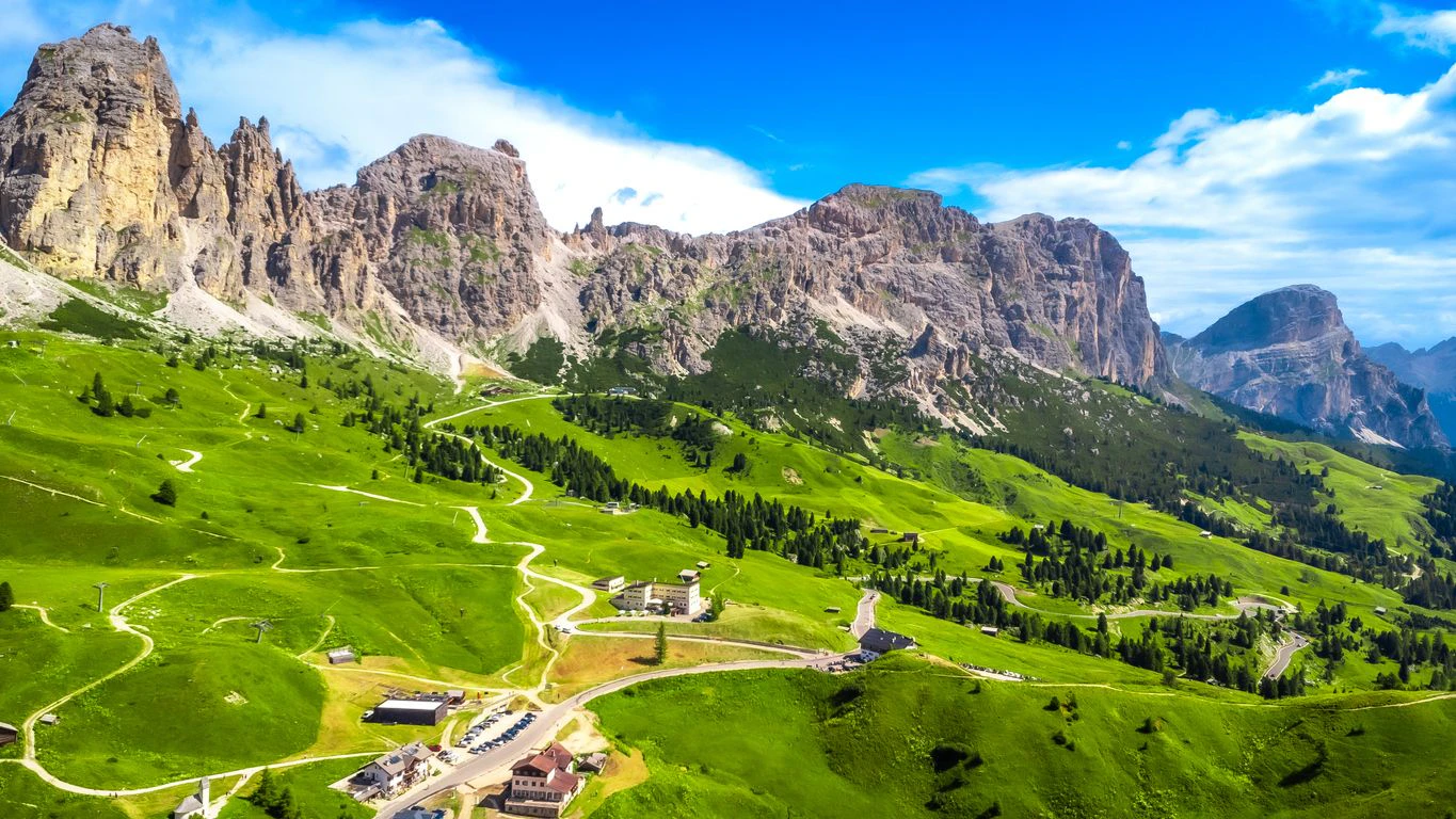 Breathtaking aerial view of the scenic gardena pass in the dolomites, showcasing winding roads, green valleys, and majestic peaks under a vibrant blue sky