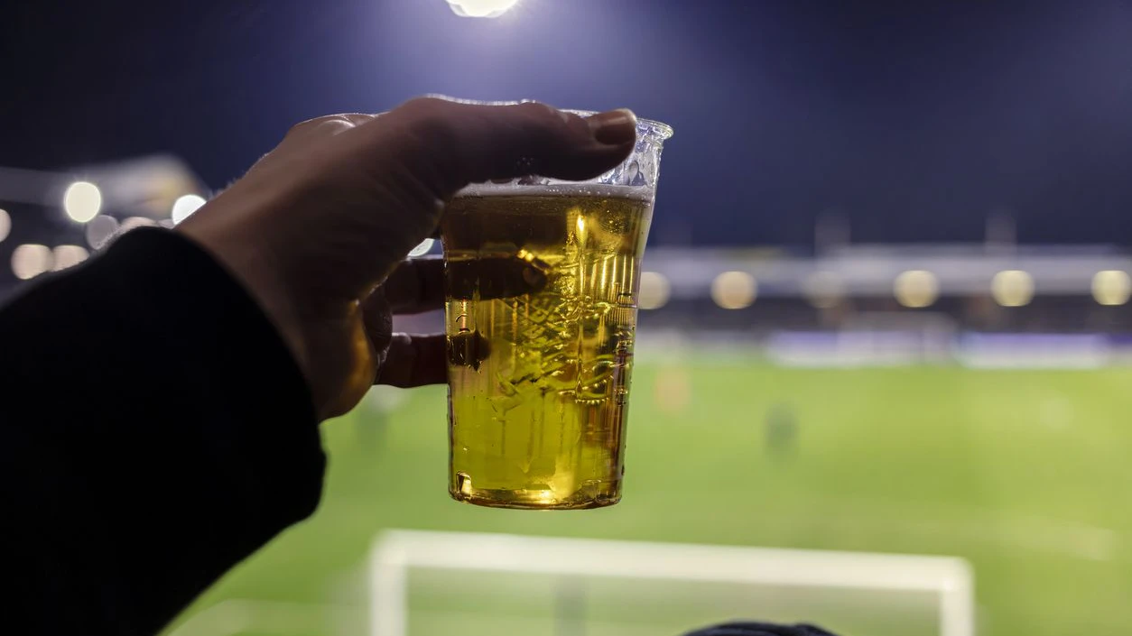Hand holding plastic cup of beer at football stadium with blurred pitch and floodlights in background, fan perspective