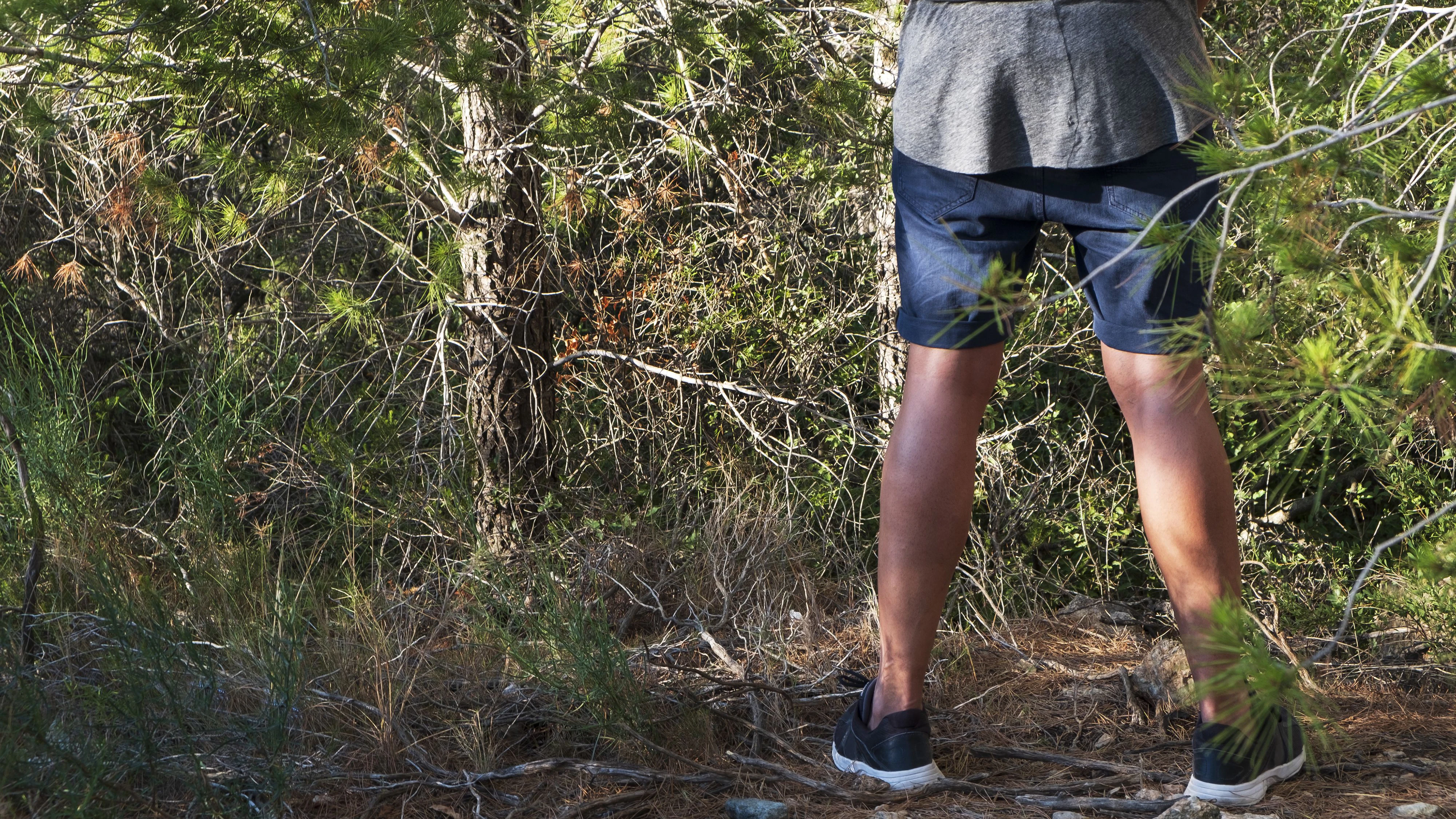 a young caucasian man wearing shorts and T-shirt seen from behind peeing in the woods