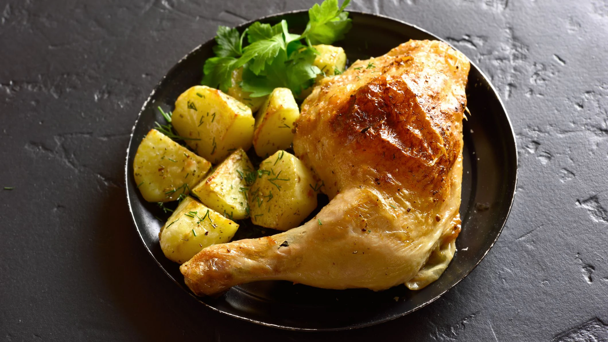 Baked chicken leg with potato on black stone background.
