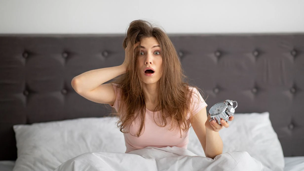 Terrified yound woman holding alarm clock in bed at home, grabbing her head, oversleeping to work. Shocked millennial lady being late, having difficulty getting up in mornings