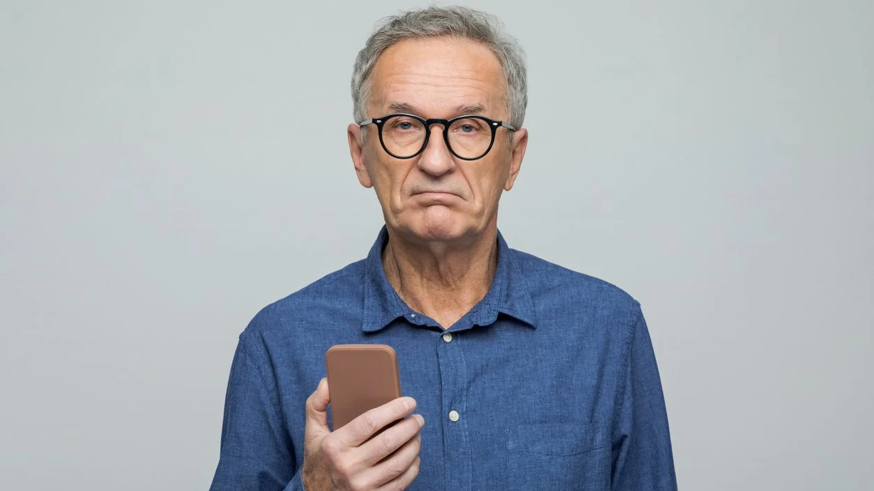 Disappointed senior man wearing denim shirt holding smart phone in hand and looking at camera, rolling eyes. Studio shot, grey background.