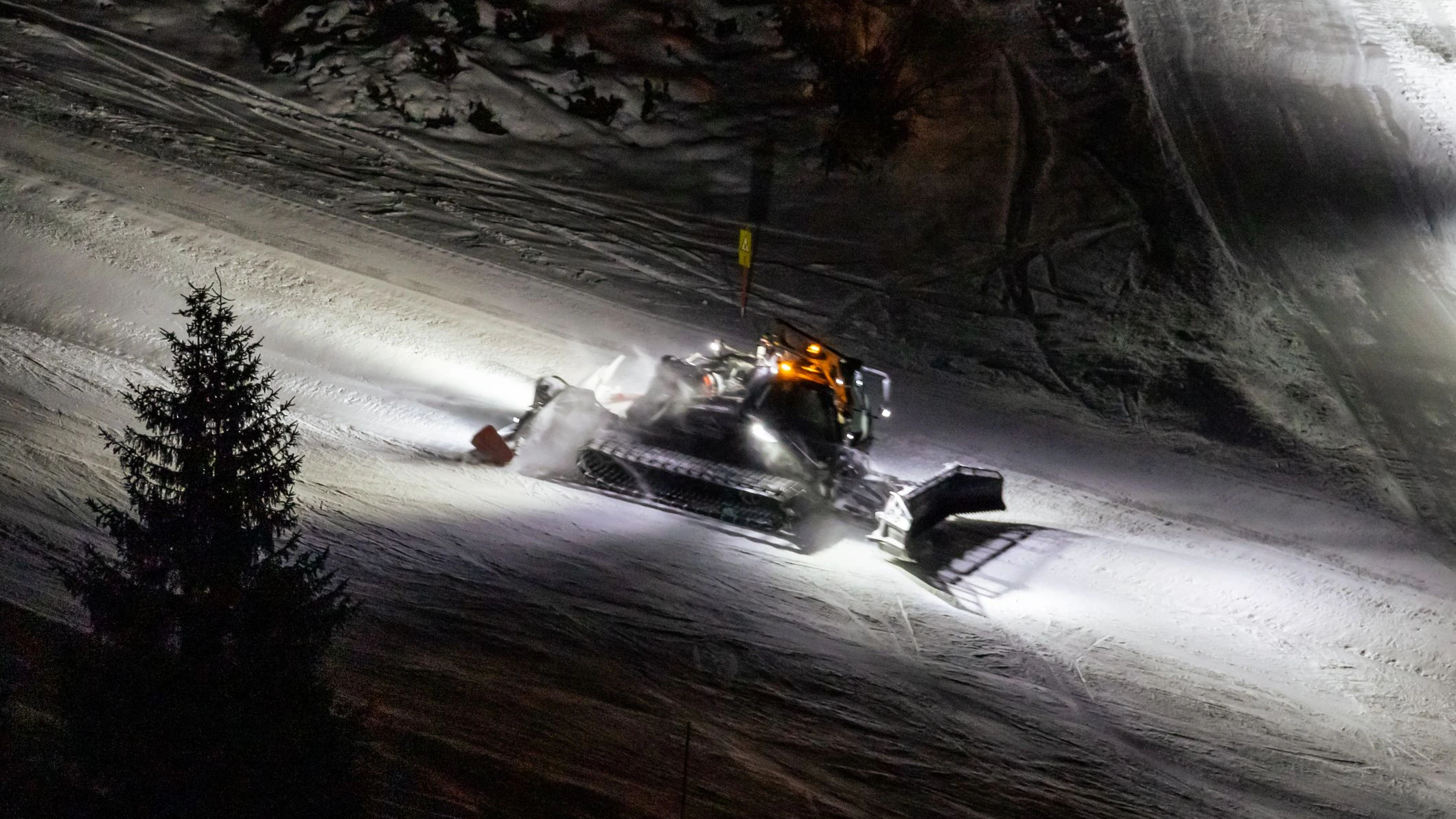 A powerful snow groomer, illuminated by bright headlights, traverses a snow-covered mountain slope at night. The machine is actively grooming the snow, leaving a smooth, prepared surface. A lone evergreen tree stands in the foreground, contrasting with the artificial light and the groomed snow. The scene evokes a sense of winter activity and the preparation for skiing or snowboarding. The photo was taken during a photowalk in Obertauern, Salzburg, Austria in December 2025.