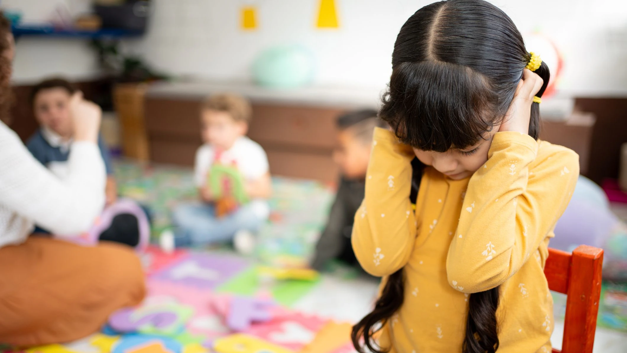 Preschool girl covers her ears and closes her eyes while other kindergarten children play loudly behind her. Concept of noise sensitivity, sensory overload, and emotional response in early childhood