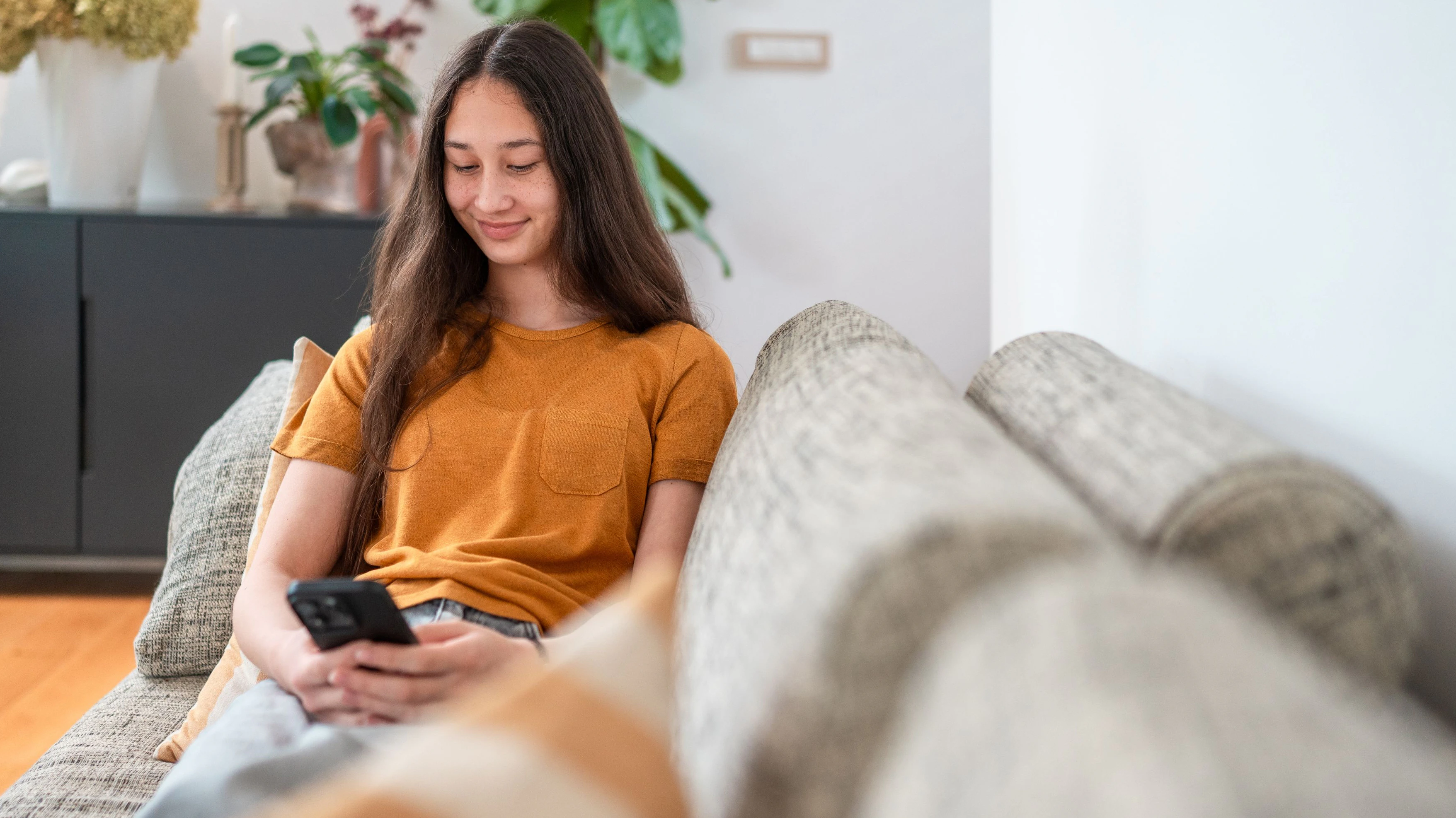 A multiracial teenage girl with long dark hair sits on a comfortable sofa, wearing a mustard shirt, using her smartphone in a bright, modern living room with plants and warm tones.