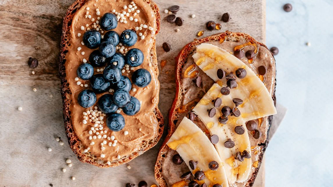 Overhead view of two toasted bread slices topped with peanut butter, almond butter, banana and blueberries