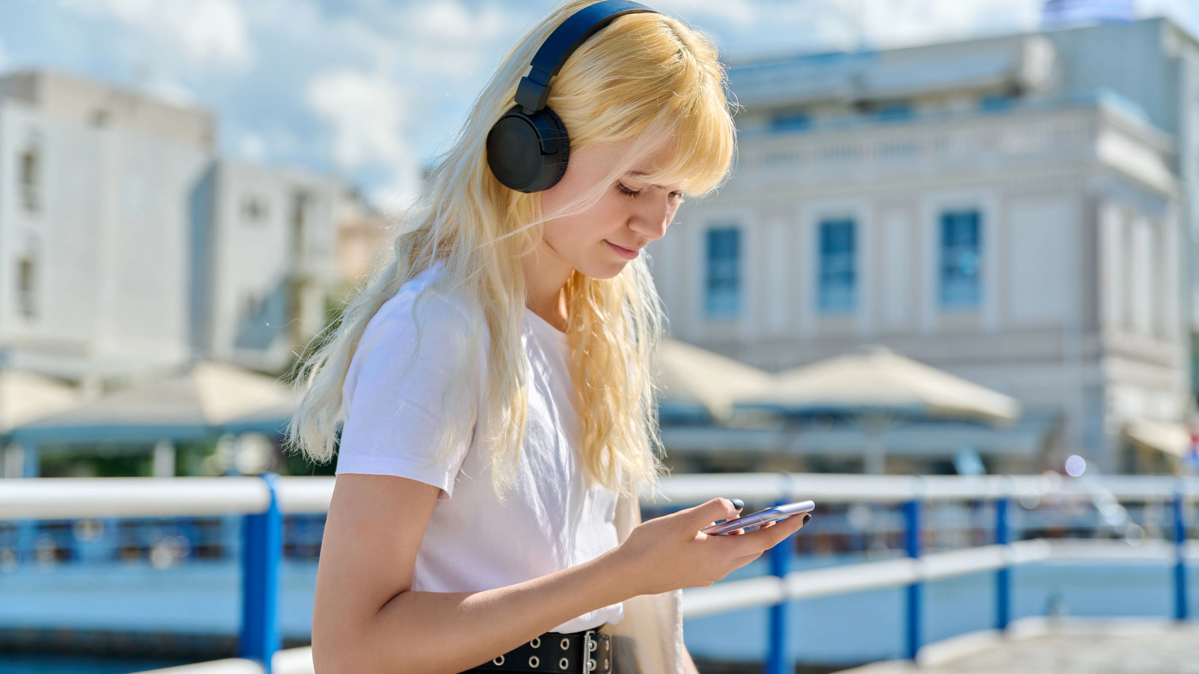 Beautiful teenage female in headphones reading looking at smartphone screen outdoor. Technology, lifestyle, adolescence, young people concept