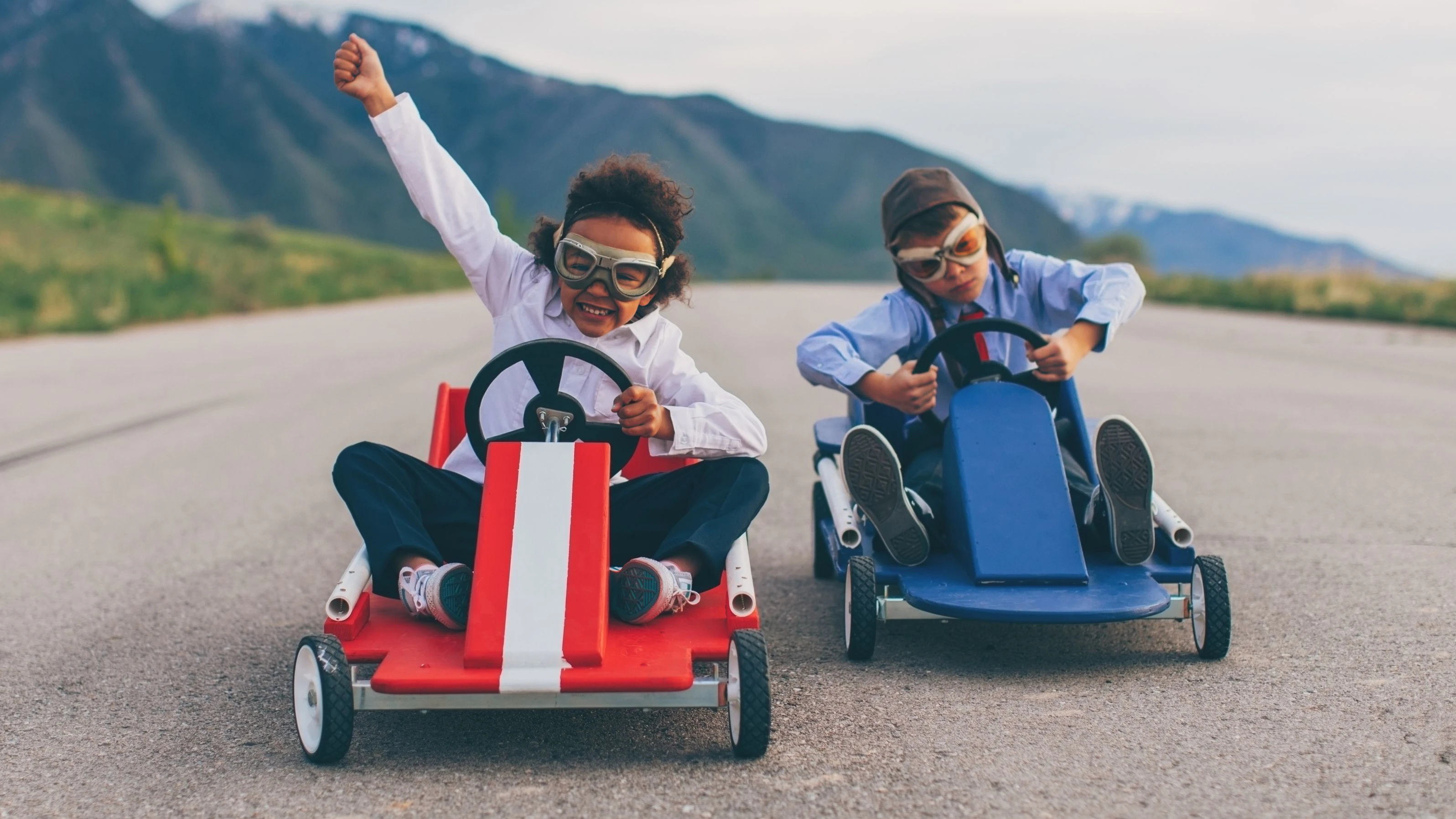 A young business girl races a boy dressed in business attire and race goggles in a push cart down a rural road in Utah. These business children love racing and competing and working hard for the success of their business. The young business boy struggles to beat the girl. The girl is winning.