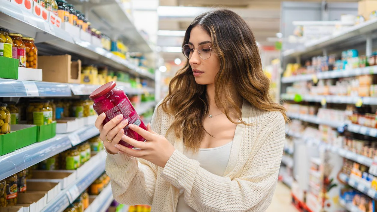 Young adult woman with glasses examining a jar in a grocery store aisle. She is focused on reading the label, surrounded by a variety of food products