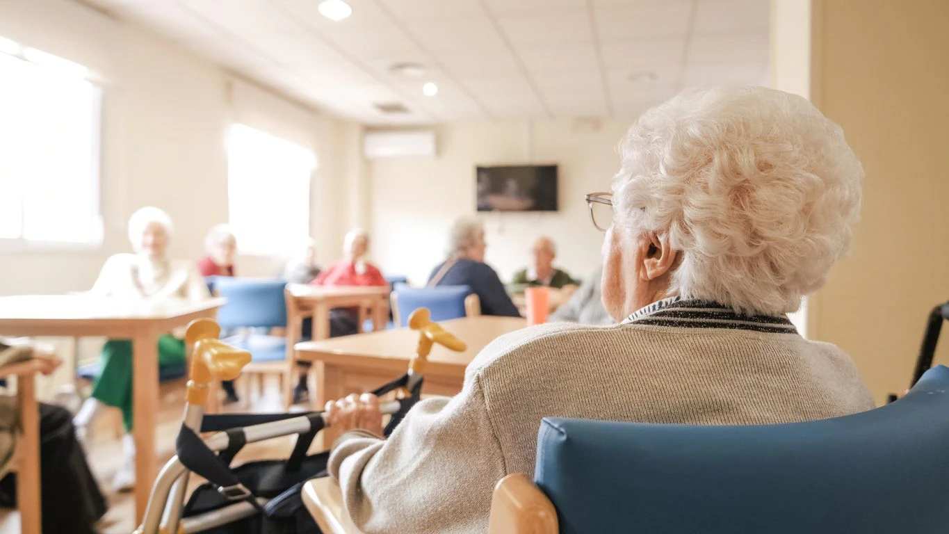 Back view of senior woman with short white hair sitting on chair with foldable walker with friends in blurred background at nursing home