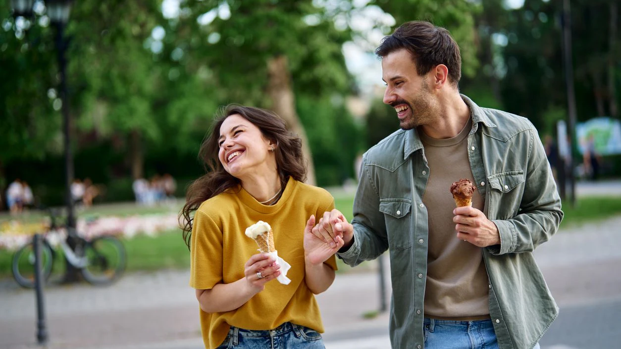 Happy young couple enjoying ice cream cones while walking hand in hand, laughing together in a city park on a bright sunny day, radiating joy and affection in their relationship
