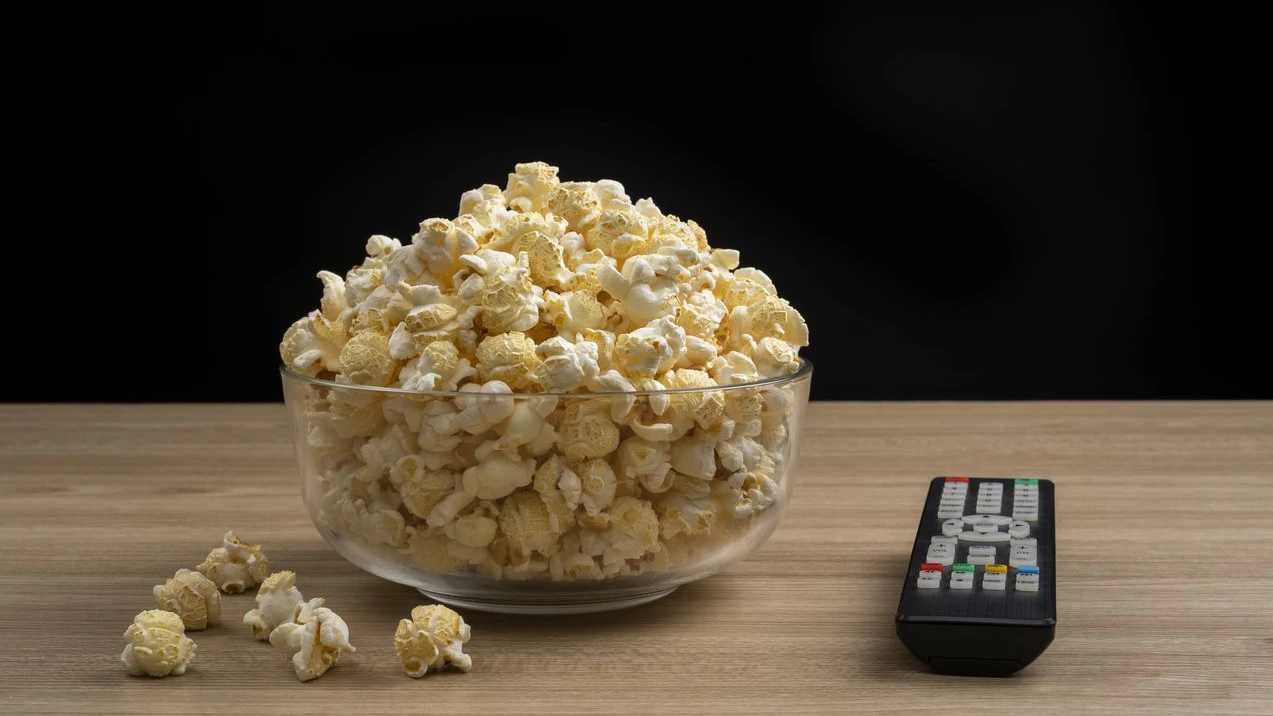 A glass bowl of popcorn and TV remote control on wooden table, black background.