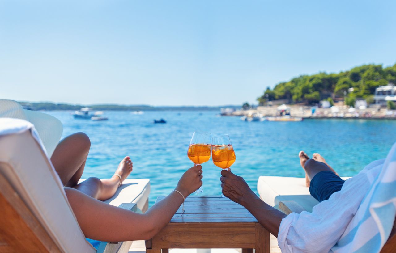 Ein Paar entspannt sich auf einer Strandterrasse mit Blick auf die kroatische Adria und stößt mit einem Spritz-Cocktail an. Das türkisfarbene Meer ist kristallklar. In der Ferne zeichnet sich der Horizont ab.