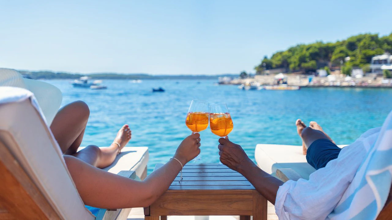 Ein Paar entspannt sich auf einer Strandterrasse mit Blick auf die kroatische Adria und stößt mit einem Spritz-Cocktail an. Das türkisfarbene Meer ist kristallklar. In der Ferne zeichnet sich der Horizont ab.