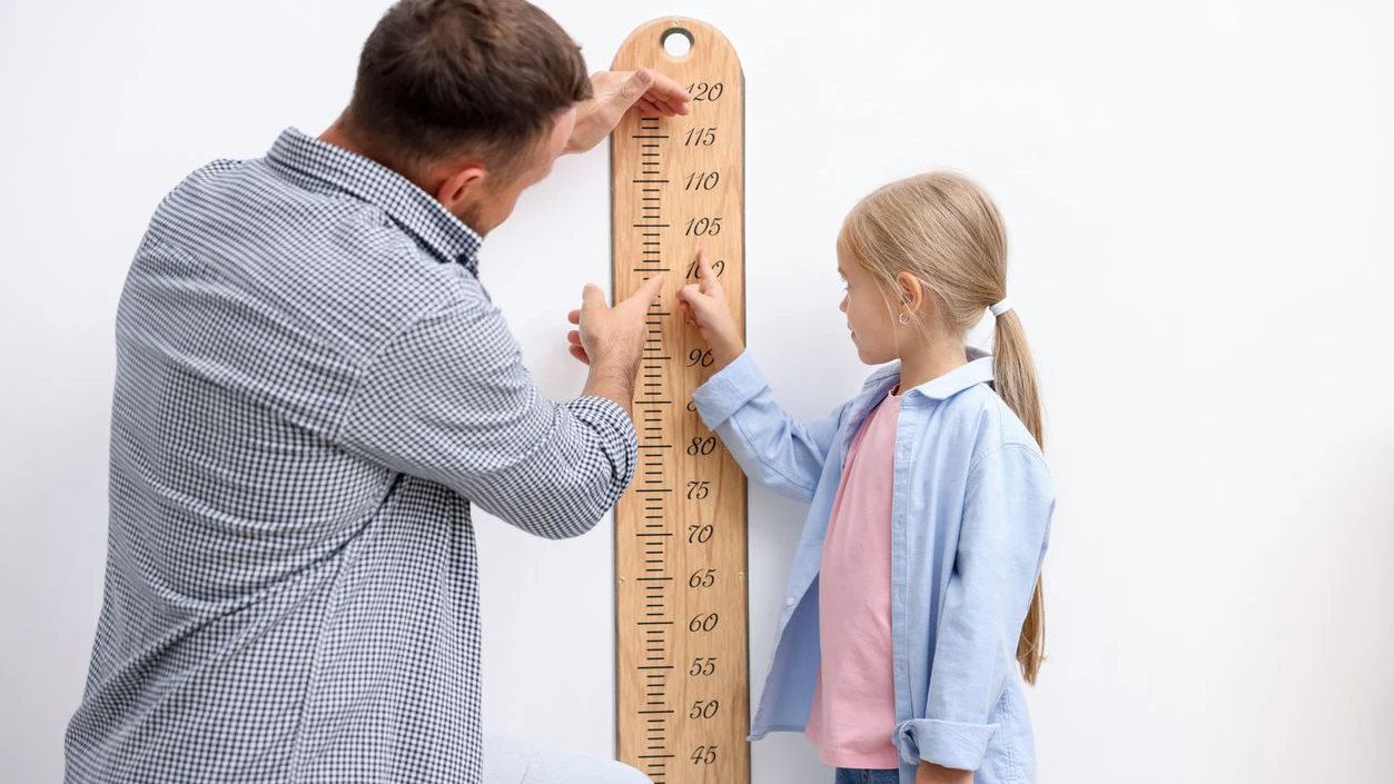 Father measuring his little daughter's height with scale near white wall indoors