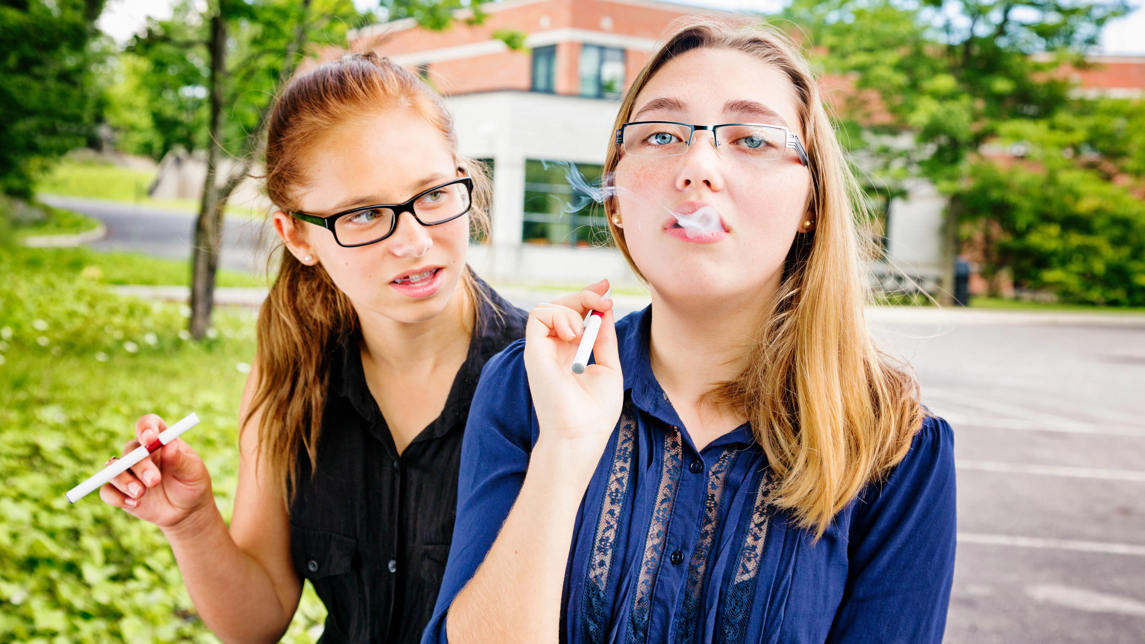 Teenage girl teaching an unsure friend how to smoke using e-cigarette. They are sitting in the high school parking lot.