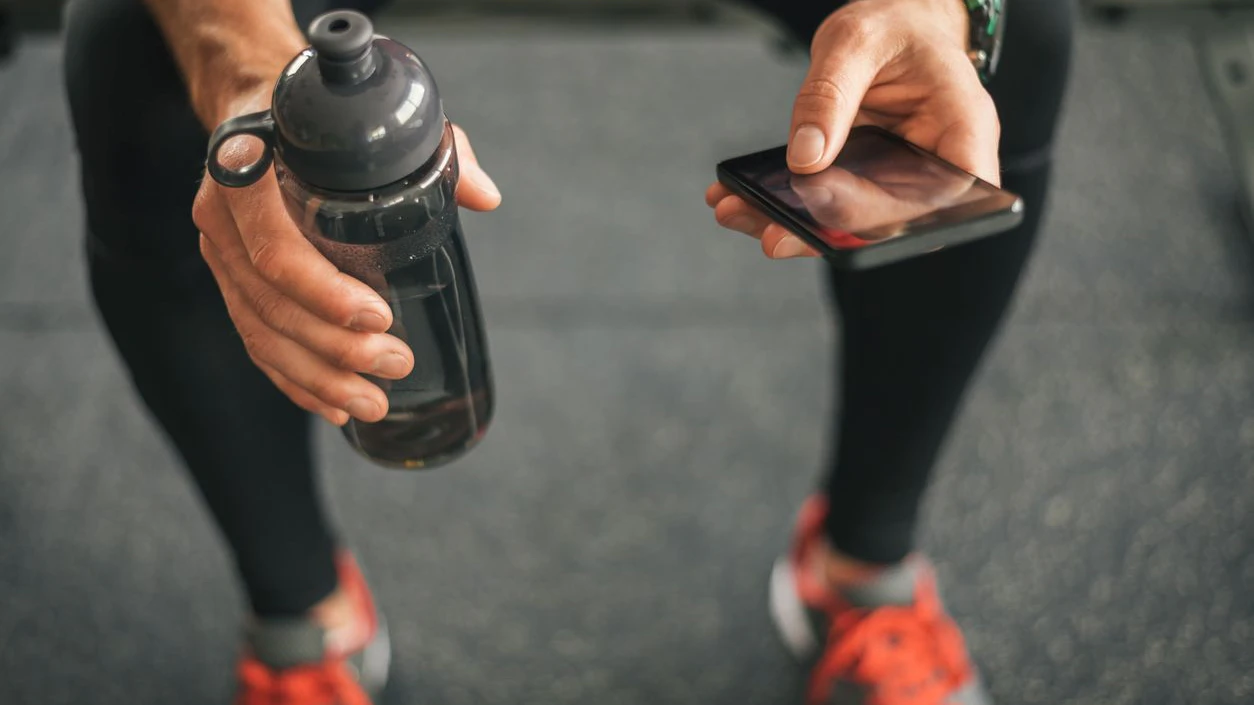 Fitness man looking to the phone for motivation before gym workout. Sporty male athlete looking his smartphone holding water bottle.
