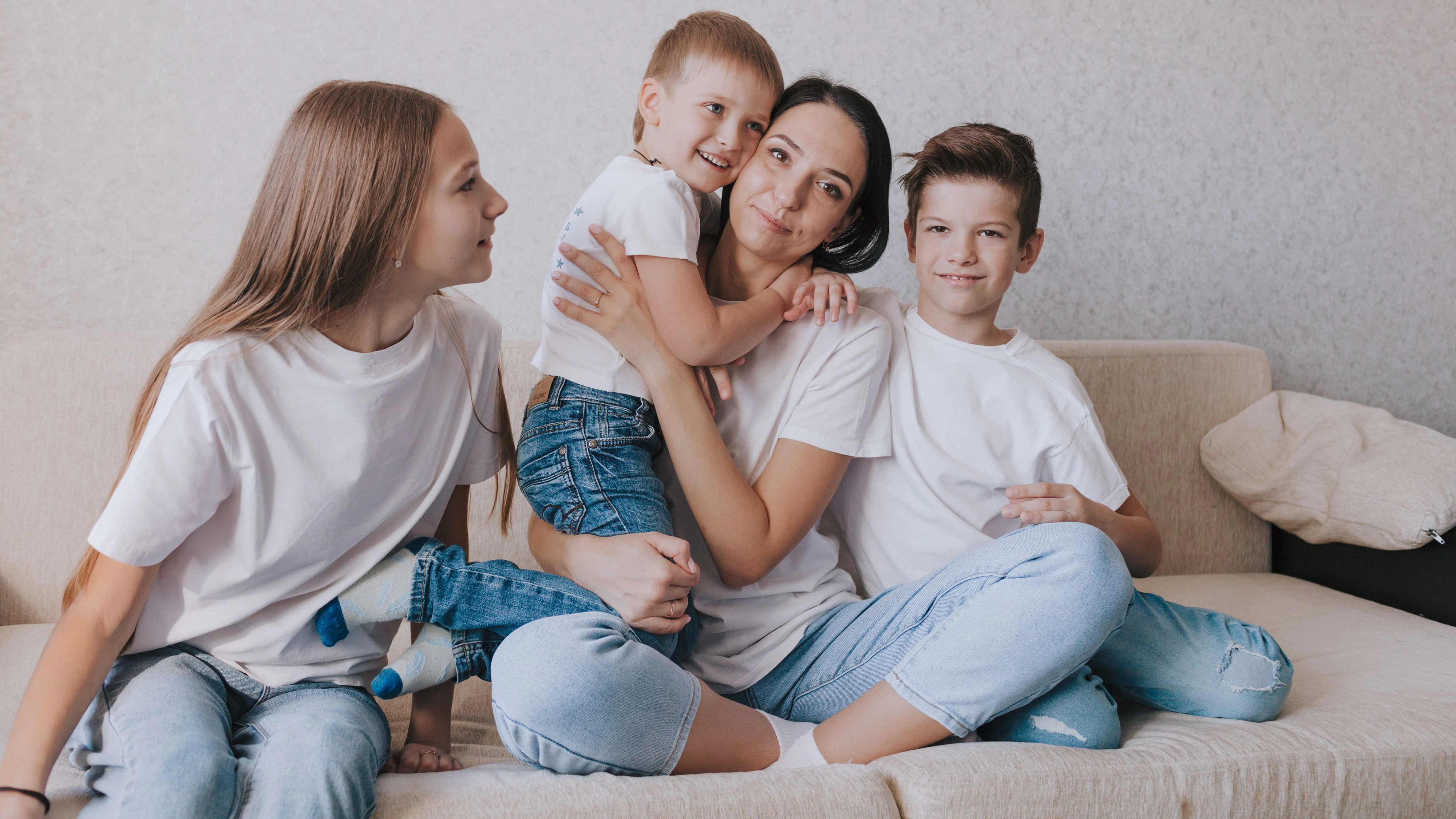 A happy family in white T-shirts and jeans is sitting on the couch. Mom hugs her children.