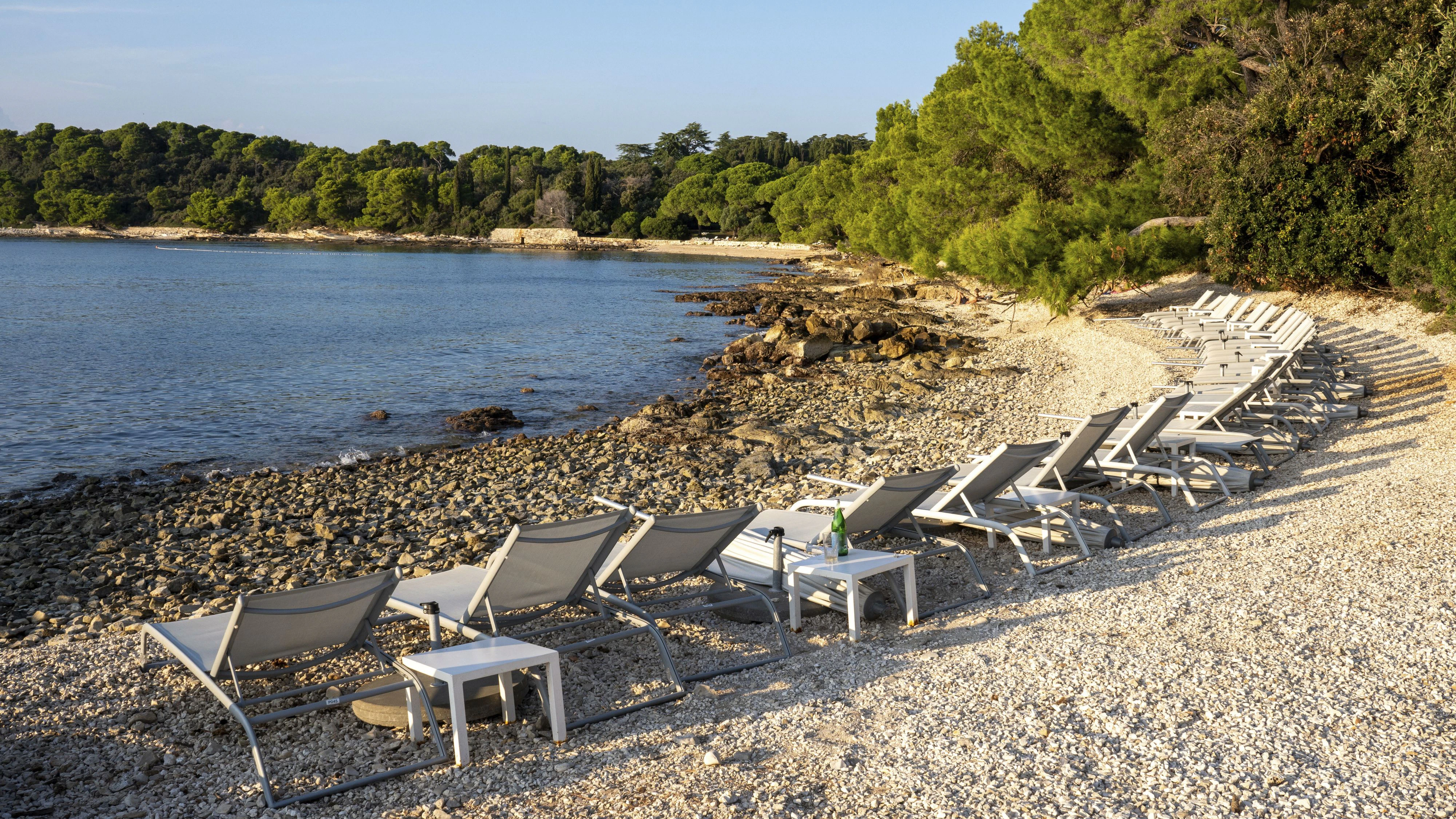 Blick auf eine Badebucht am Kieselstrand mit Liegestühlen in der Abendsonne an der Mittelmeerküste in Kroatien. Im Hintergrund Kiefernwald