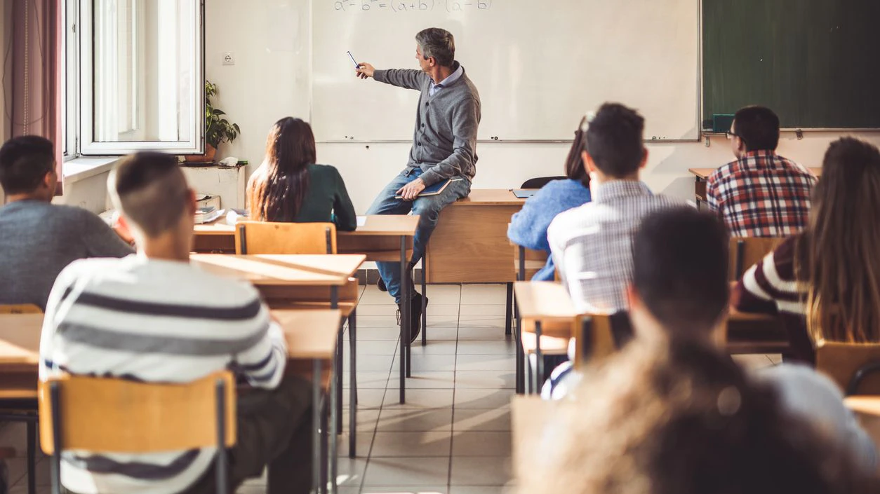 Male professor sitting on the table and teaching mathematics from whiteboard at high school.