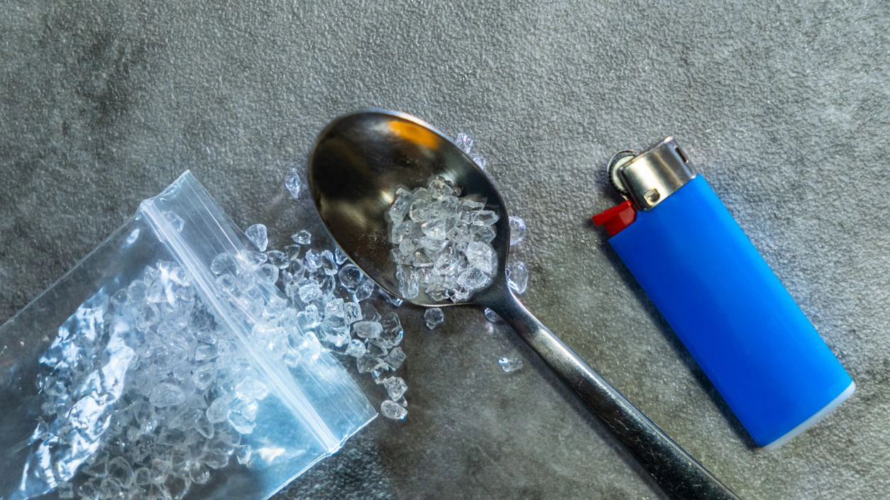 Crystal meth, blue cigarette lighter, and burnt spoon arranged on concrete floor, representing illegal drug use and addiction themes.