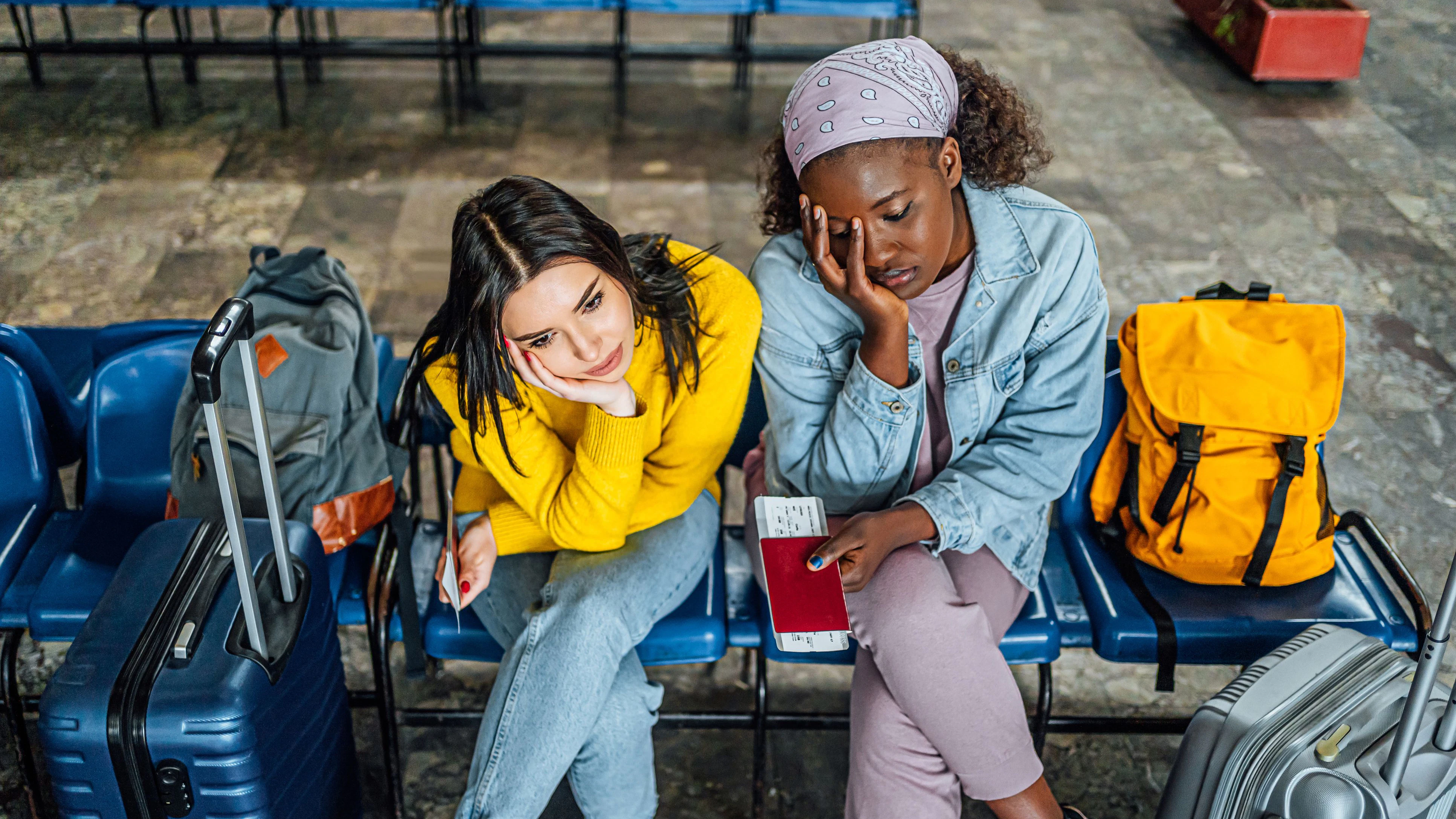 Young multi racial women friends with suitcases sitting bored, waiting for a train at railway station