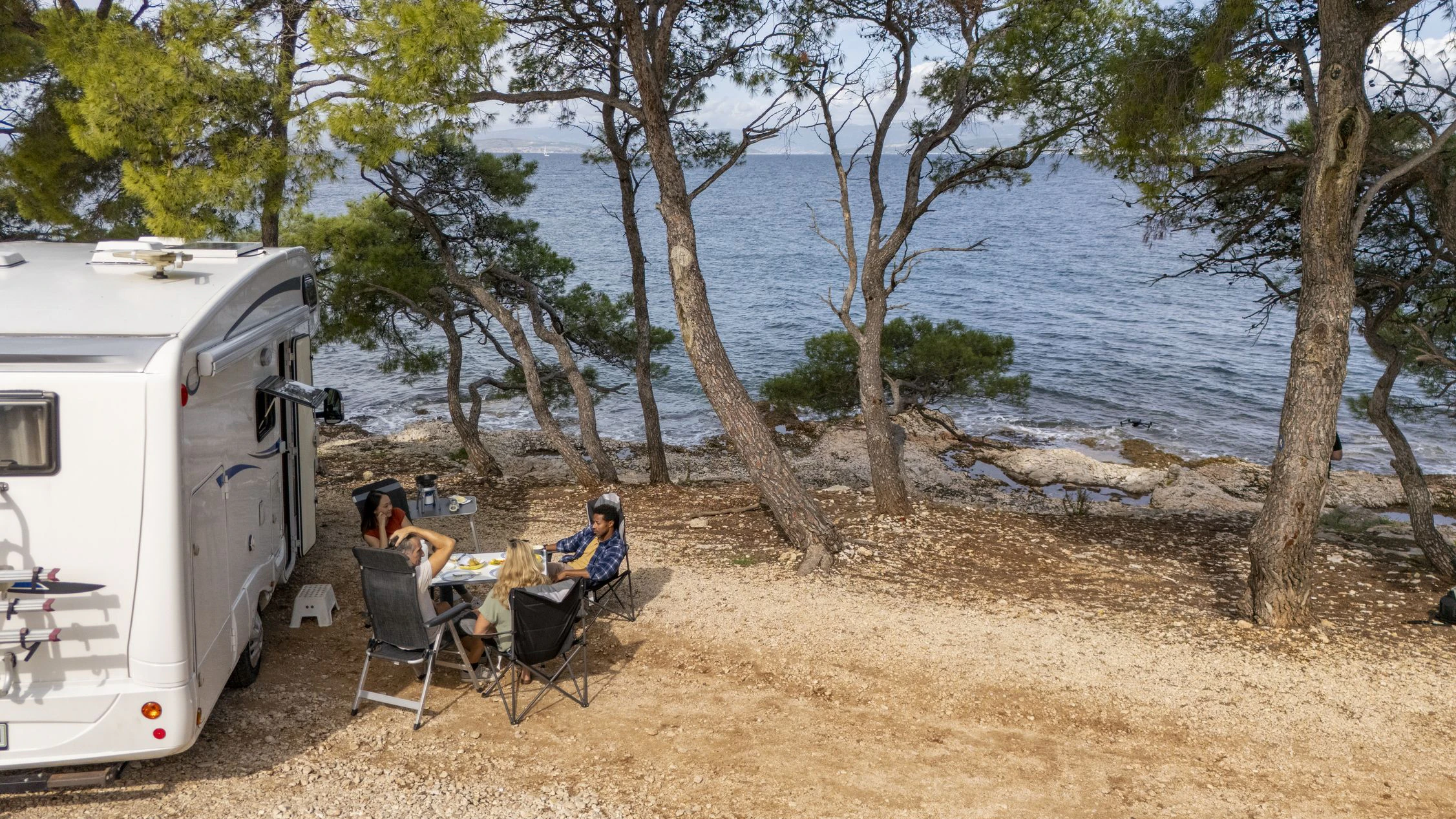 Diverse group of friends relaxing outside a camper by the sea, surrounded by pine trees, under bright daylight with a calm coastal breeze. Camping Campingurlaub Kroatien Meer Wohnwagen