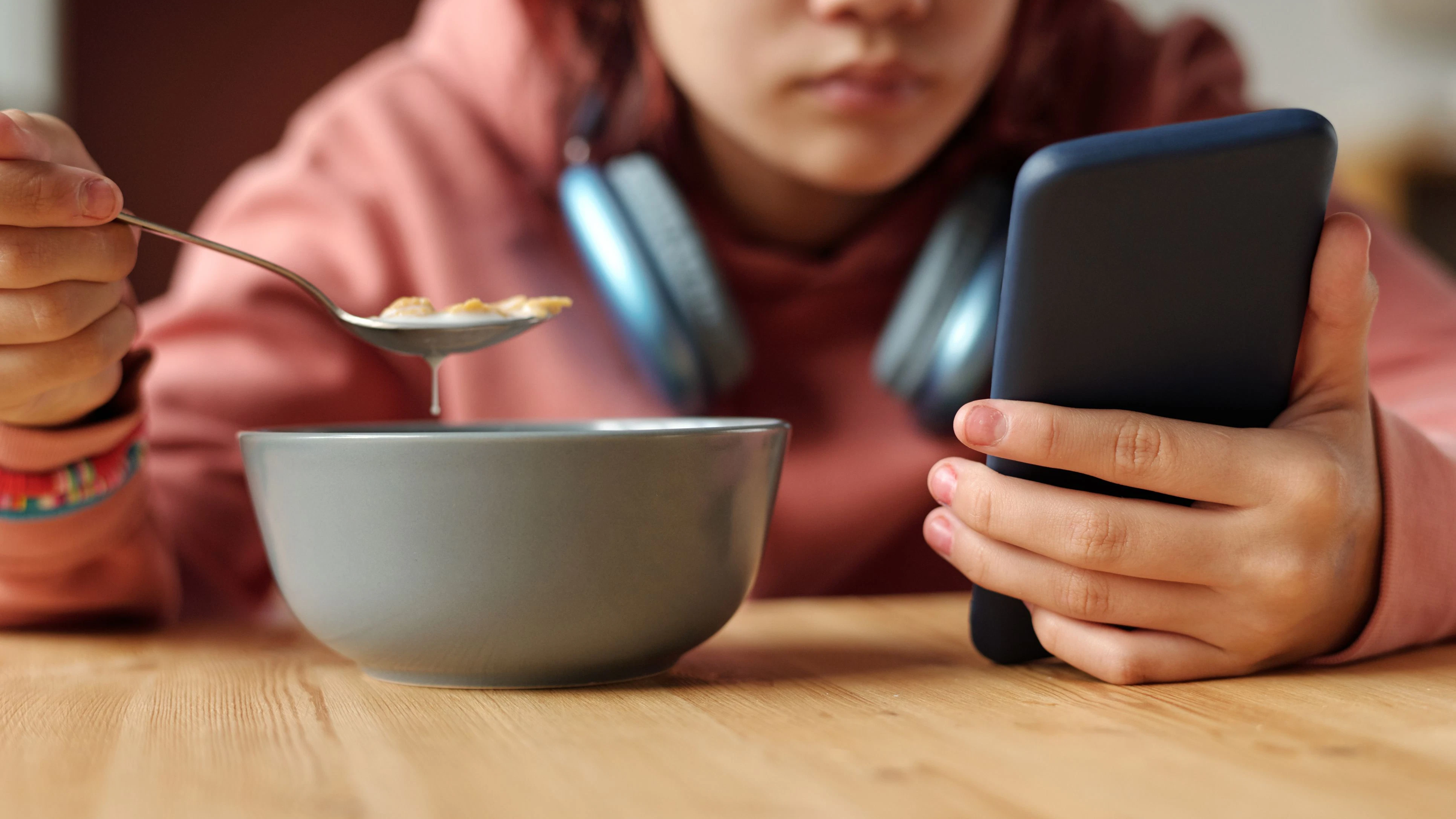 Hand of teenage girl with mobile phone having cornflakes with milk from bowl while sitting by table in front of camera and reading messages