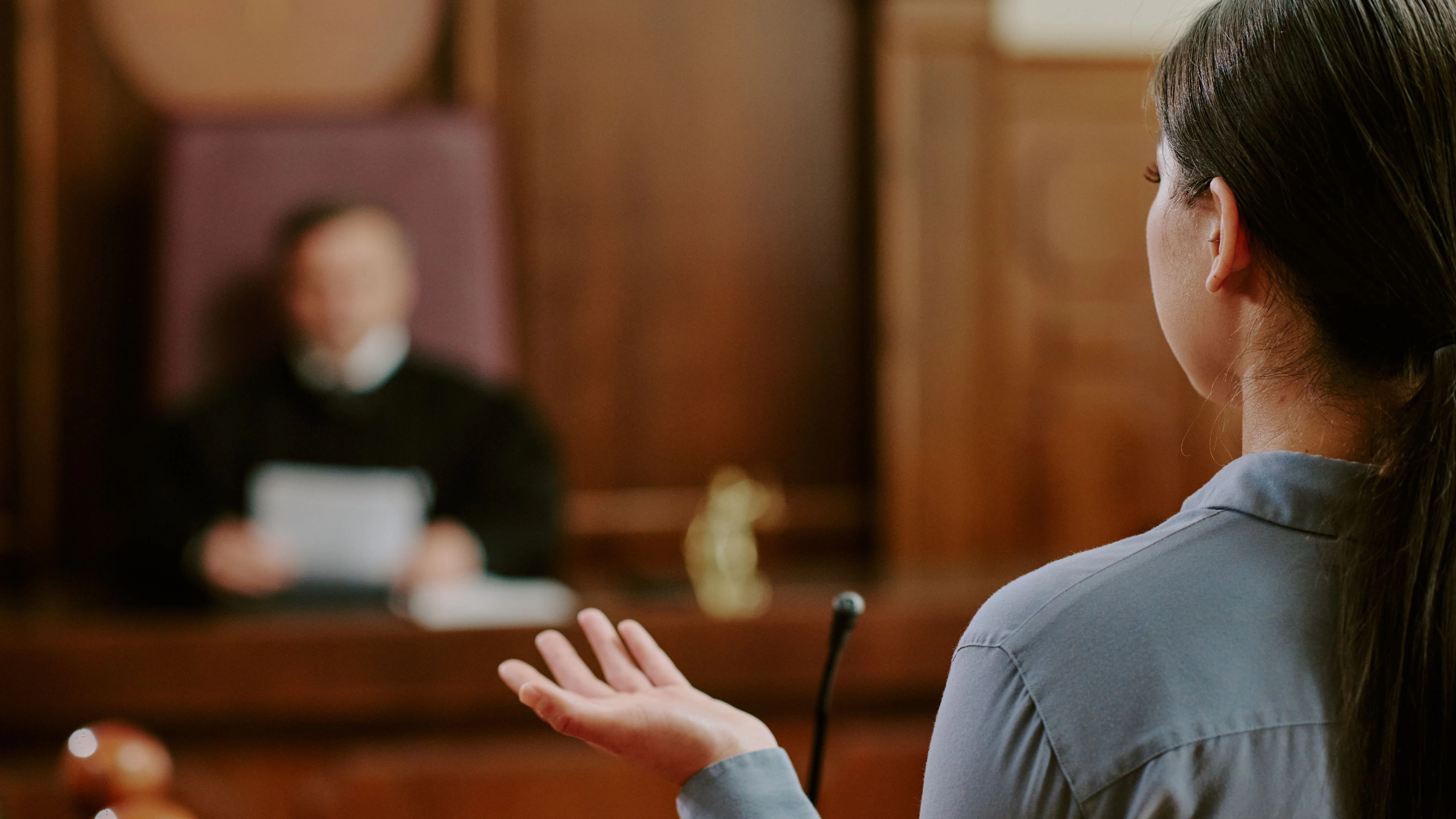 Young adult Asian woman testifying in courtroom, standing in front of judge, gesturing with hand while speaking, legal proceeding taking place in judicial setting