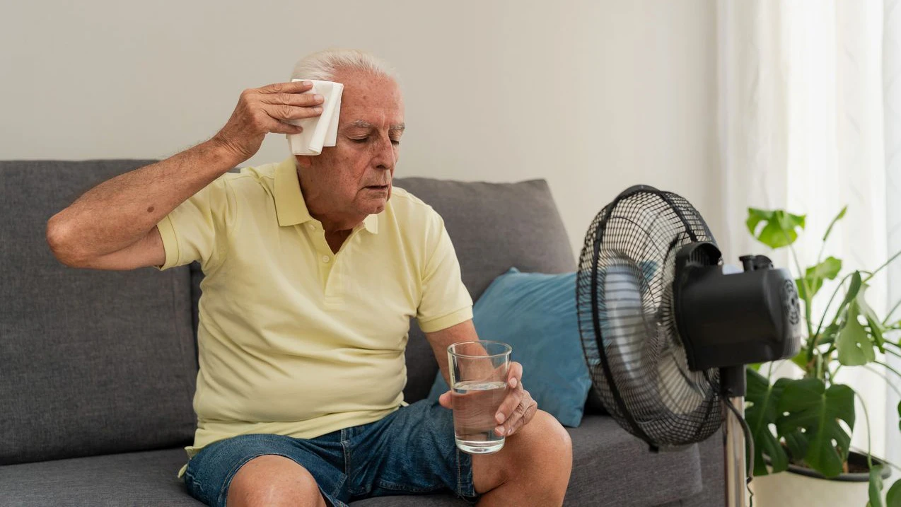 Senior man sitting on sofa wiping sweat from forehead while holding glass of water near ventilator