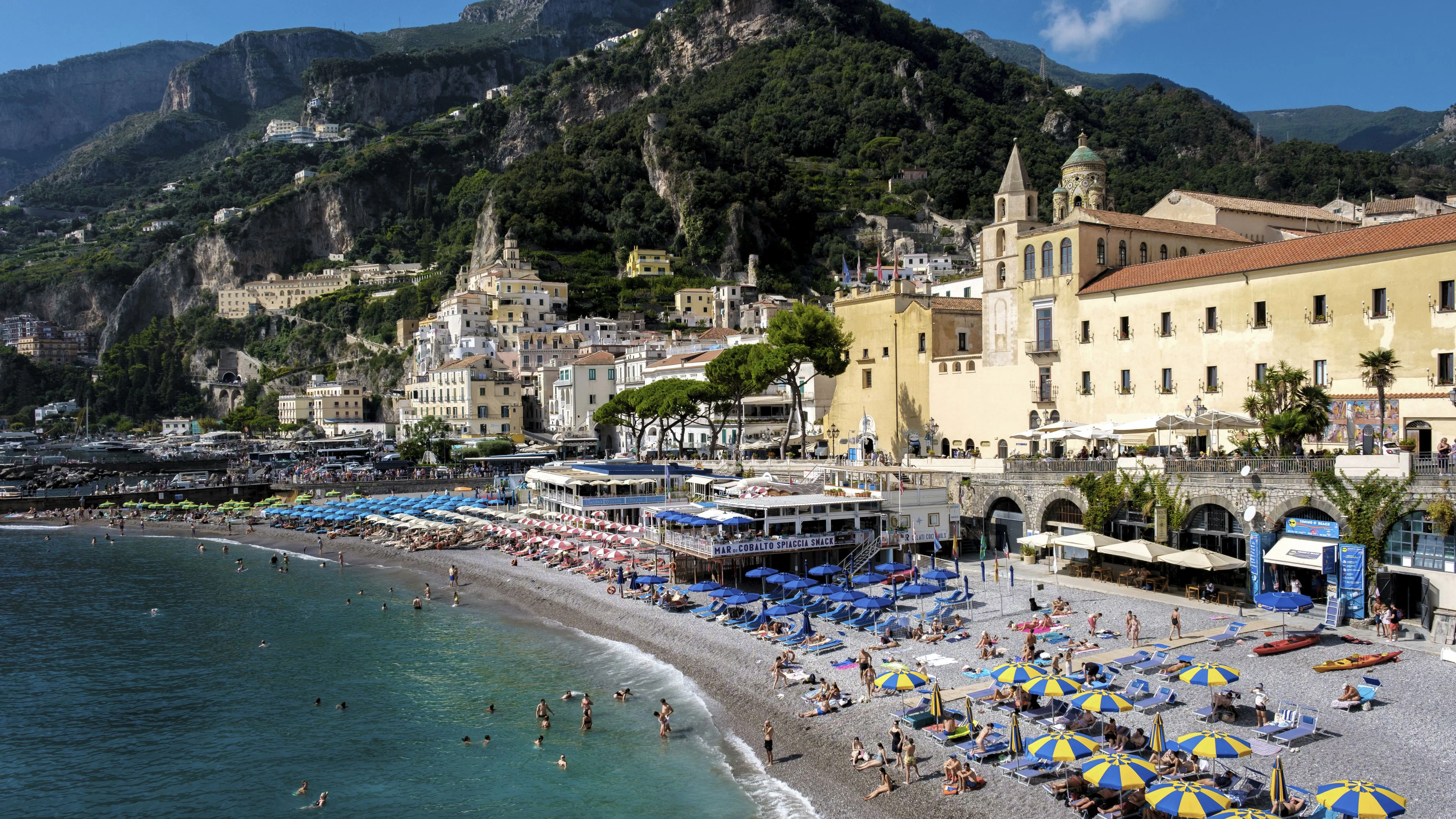 Tourists bask under colorful umbrellas on a pebbled beach as the sun brightens the Amalfi Coast. Scenic cliffs and charming buildings create a stunning backdrop to this Mediterranean getaway.