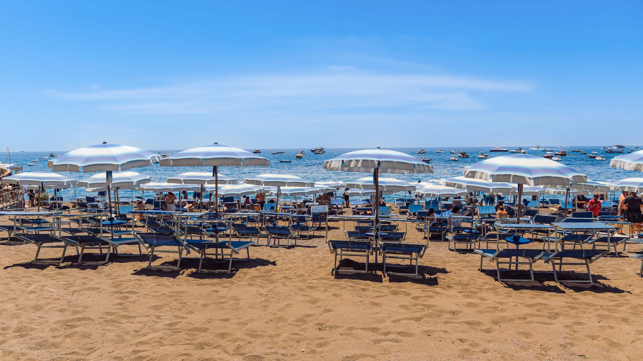 Positano, Italy - June 15 2024: sun umbrellas and lounge chairs line the sandy beach in Amalfi Coast, while boats drift in the sparkling blue water under a clear sky.