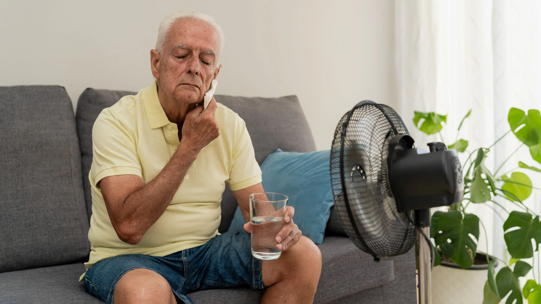 Senior man sitting on sofa wiping sweat from face holding glass of water near electric fan at home
