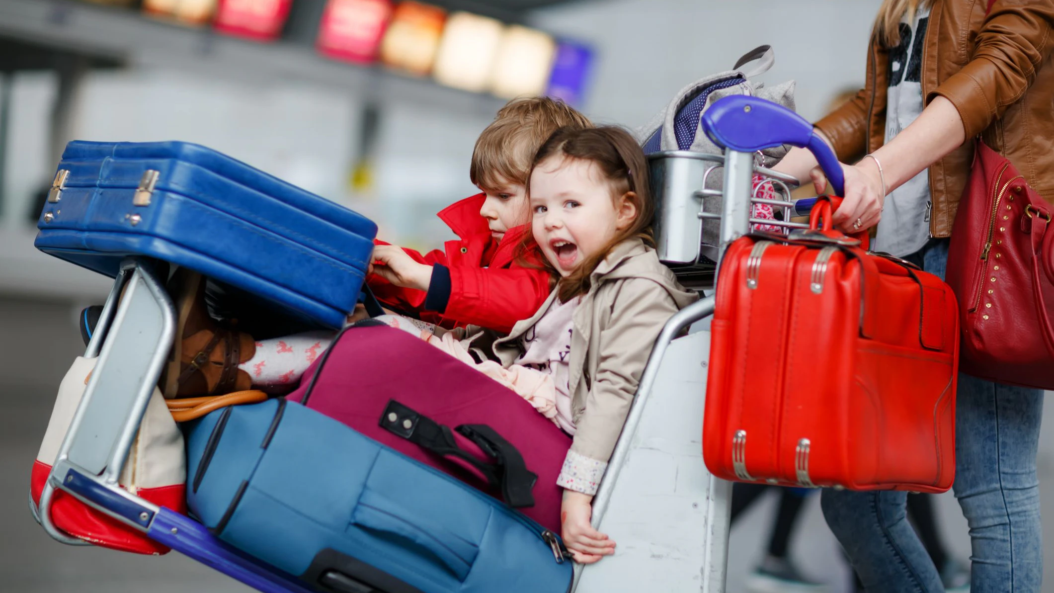 Two little kids, boy and girl, siblings and mother at the airport. Children, family traveling, going on vacation by plane and waiting on trolley with suitcases pushing by woman at terminal for flight