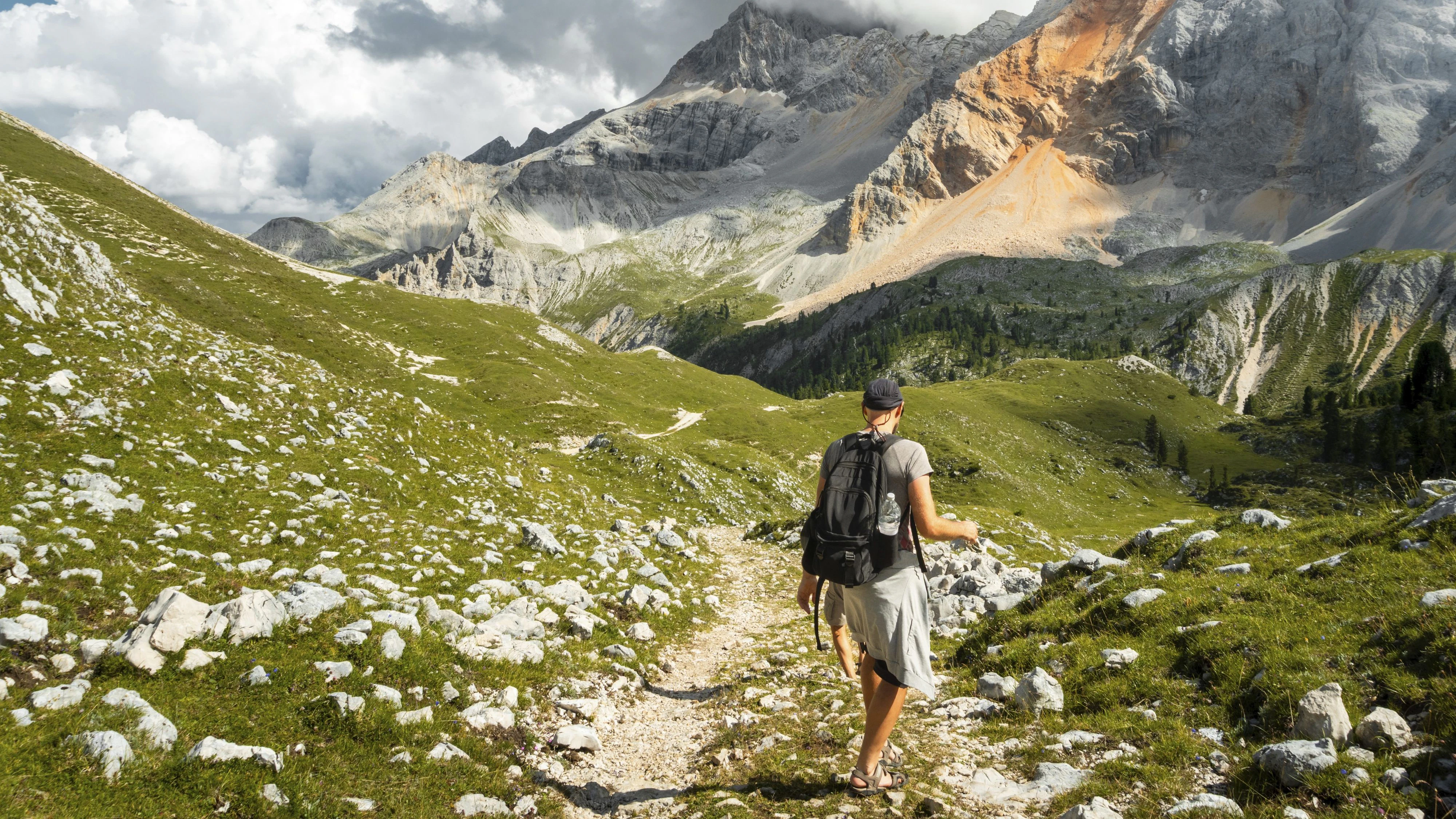 Male hiker with backpack walking dirt trail green alpine meadow majestic mountain peaks background Malga Rossalm Dolomites Italy summer adventure travel lifestyle