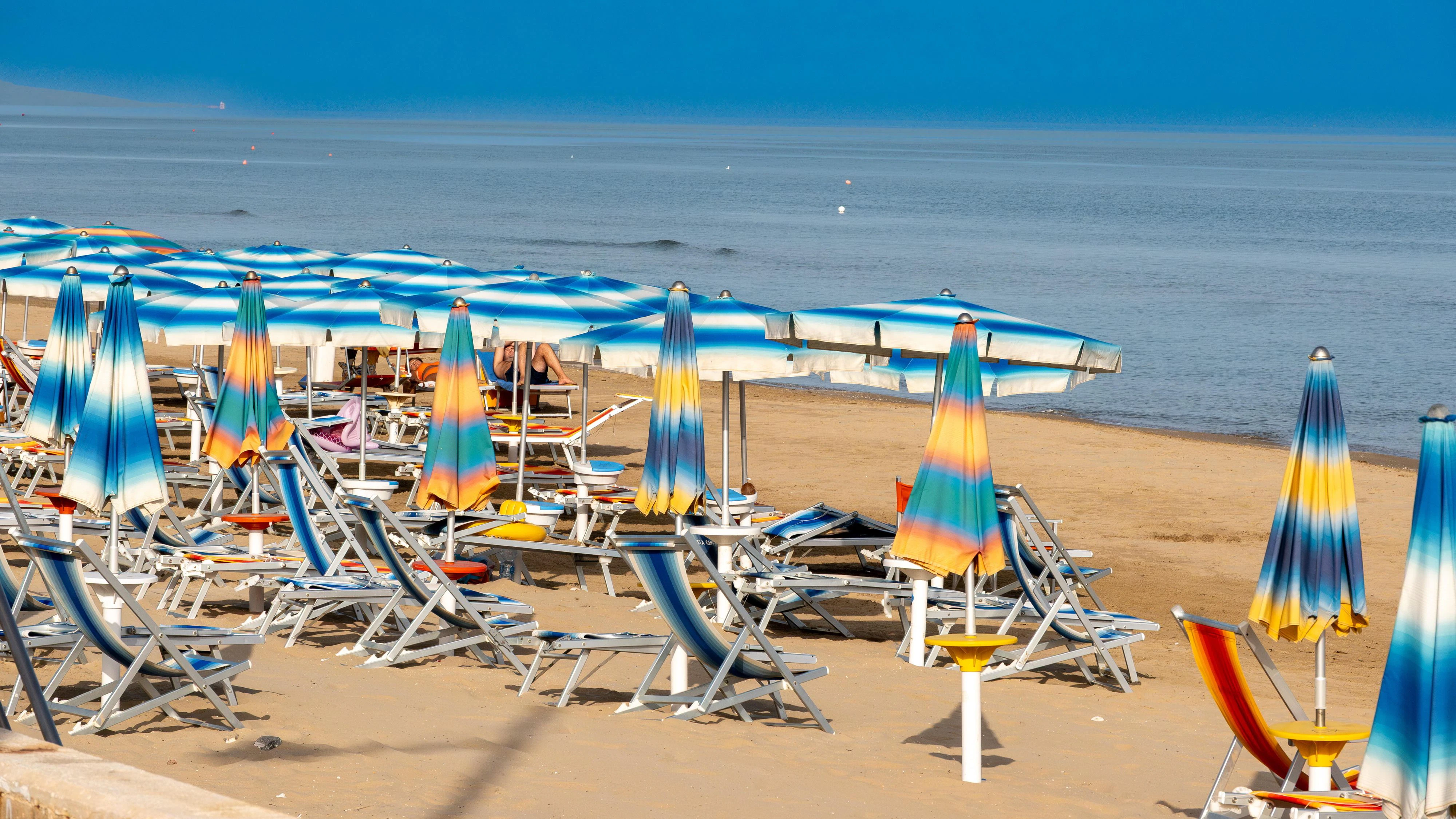 Foce Varano - Apulia - Italy - 08-28-2025: Blue and white beach umbrellas shade sun loungers on the sandy shore beside a calm sea, with a distant hazy coastline under clear sky.