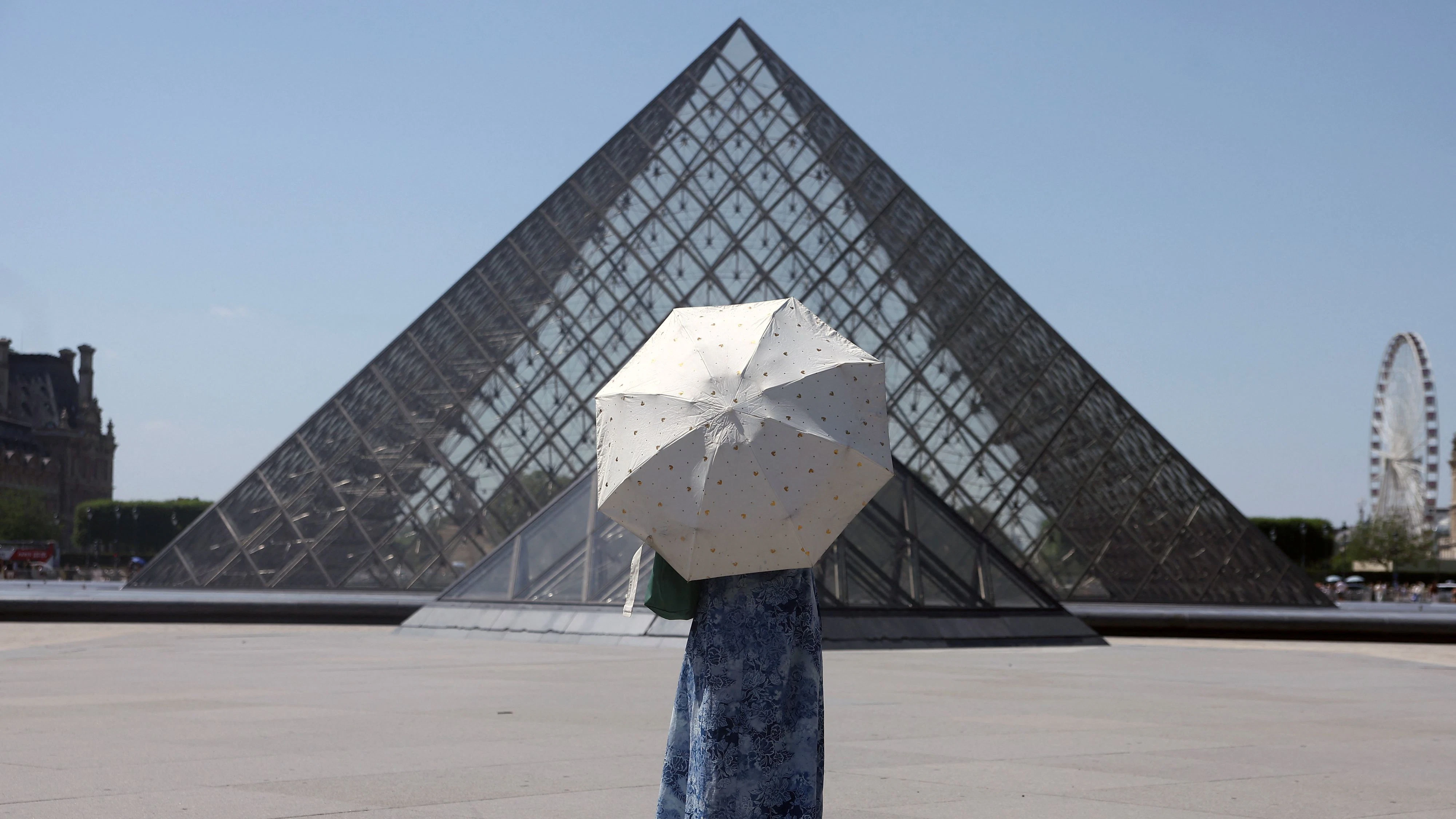 A tourist protecting herself from the sun with an umbrella stands in front of the glass Pyramid of the Louvre museum as the museum closes early due to the heatwave as temperatures rise in Paris, France, June 24, 2026. REUTERS/Alice Sacco 