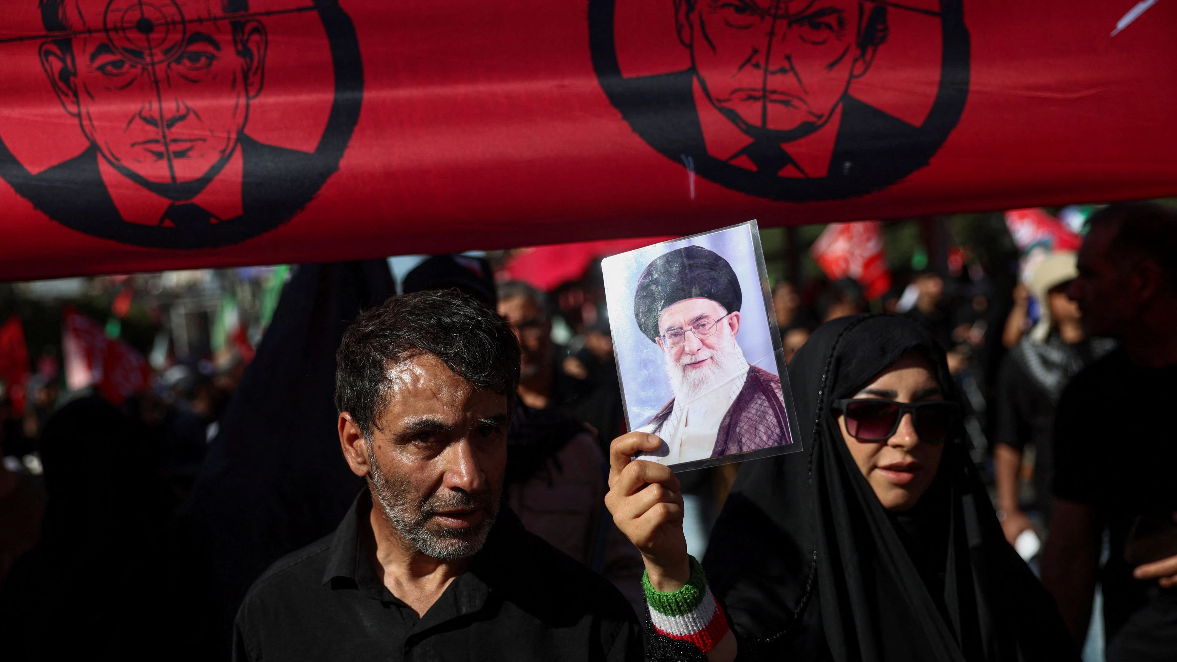 A woman holds a picture of Iran's late Supreme Leader Ayatollah Ali Khamenei as she stands underneath a banner against U.S. President Donald Trump and Israeli Prime Minister Benjamin Netanyahu as mourners gather on the day of the burial of Iran's late Supreme Leader, who was killed on February 28 in Israeli and U.S. airstrikes, in a culmination of a mass funeral, in Mashhad, Iran, July 9, 2026. Majid Asgaripour/WANA (West Asia News Agency) via REUTERS ATTENTION EDITORS - THIS PICTURE WAS PROVIDED BY A THIRD PARTY. ISRAEL OUT. NO COMMERCIAL OR EDITORIAL SALES IN ISRAEL. NO ACCESS FOR ISRAELI MEDIA. NO USE BBC PERSIAN. NO USE VOA PERSIAN. NO USE MANOTO. NO USE IRAN INTERNATIONAL. NO USE RADIO FARDA. DIGITAL: NO USE BBC PERSIAN. NO USE VOA PERSIAN. NO USE MANOTO. NO USE IRAN INTERNATIONAL. NO USE RADIO FARDA