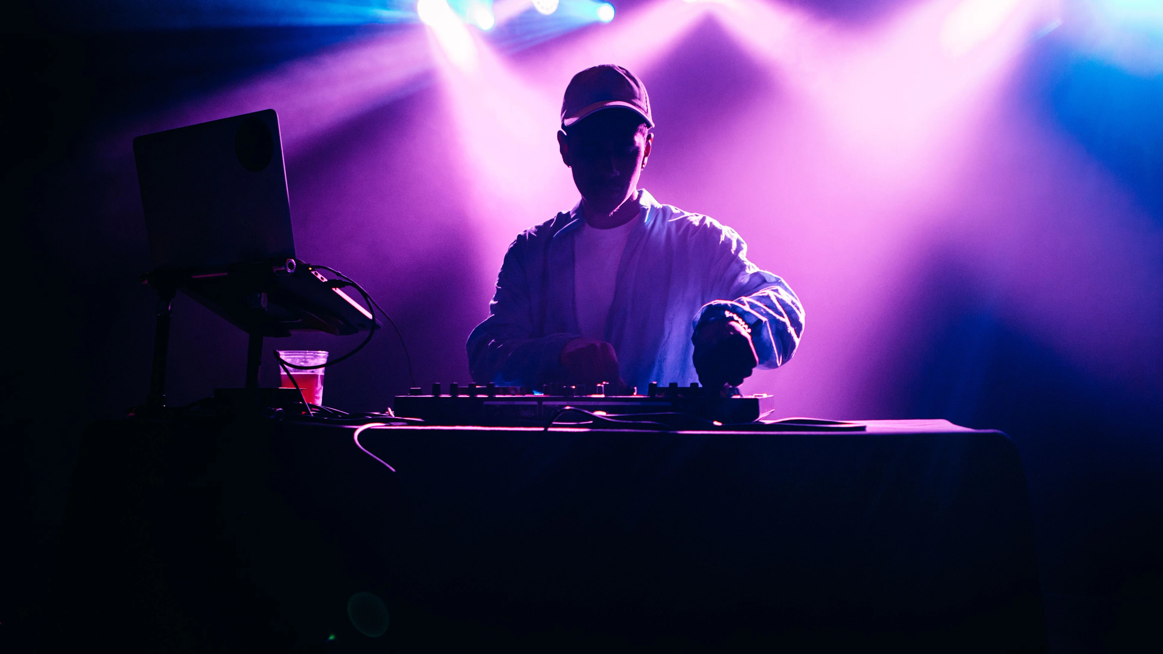 A young African American deejay performs for a crowd at a city night club. Colorful stage lights illuminate the stage behind him.