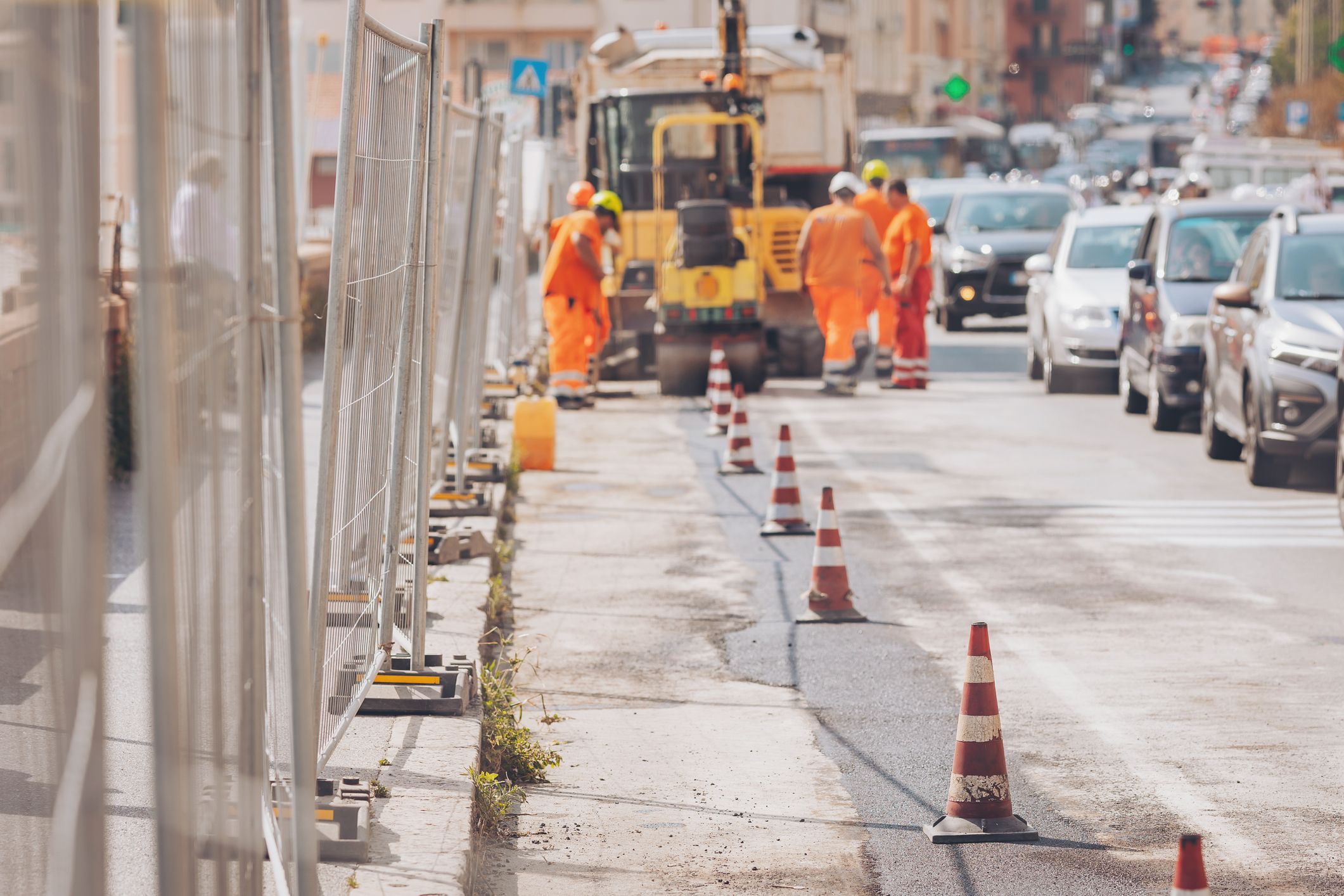 Bauarbeiter in leuchtend orangefarbener Schutzkleidung reparieren die Straße, während Absperrungen den Verkehr um die Baustelle herumleiten. 