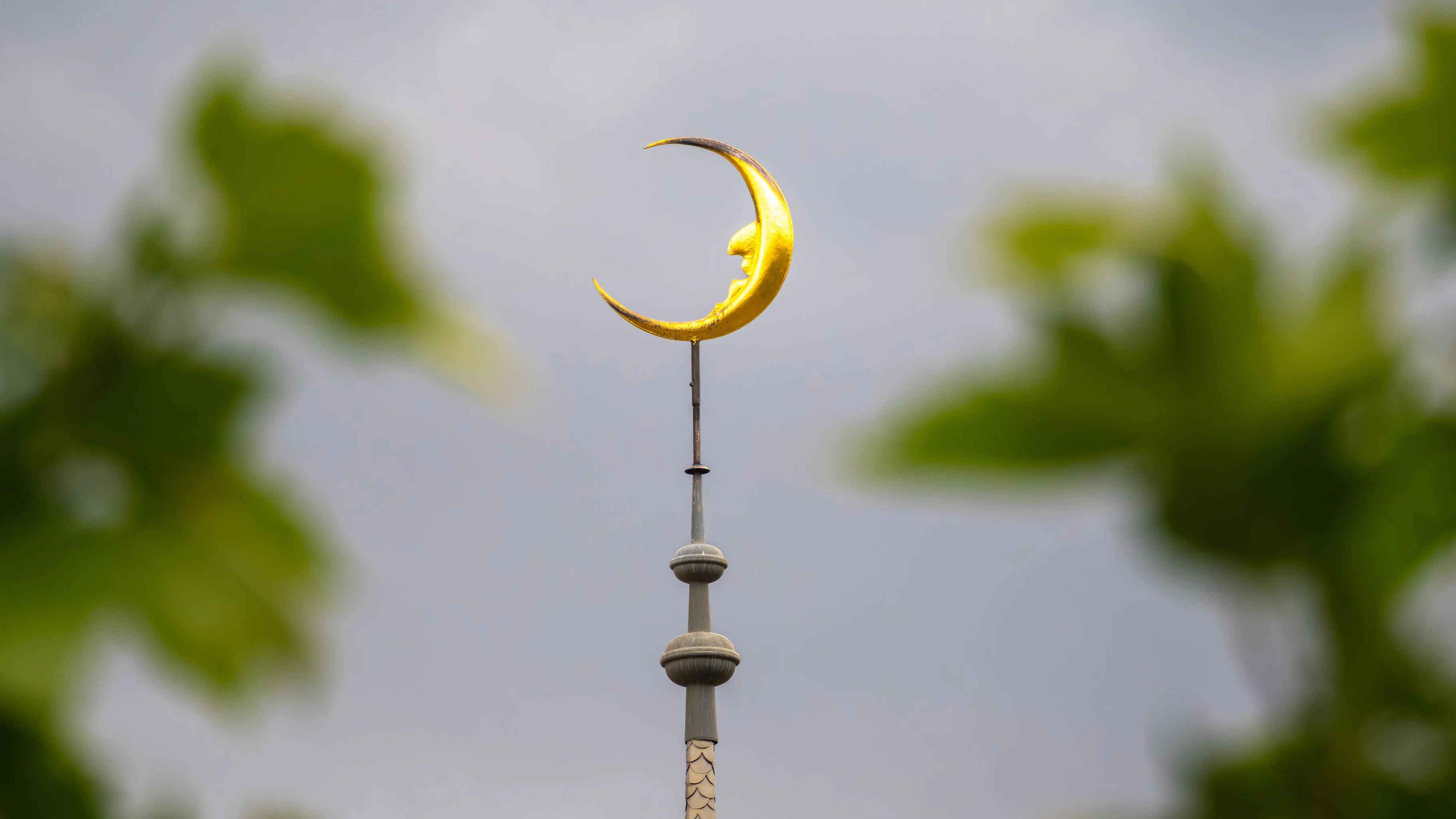 A golden crescent moon atop a building tower, framed by blurred green foliage, against a cloudy sky.