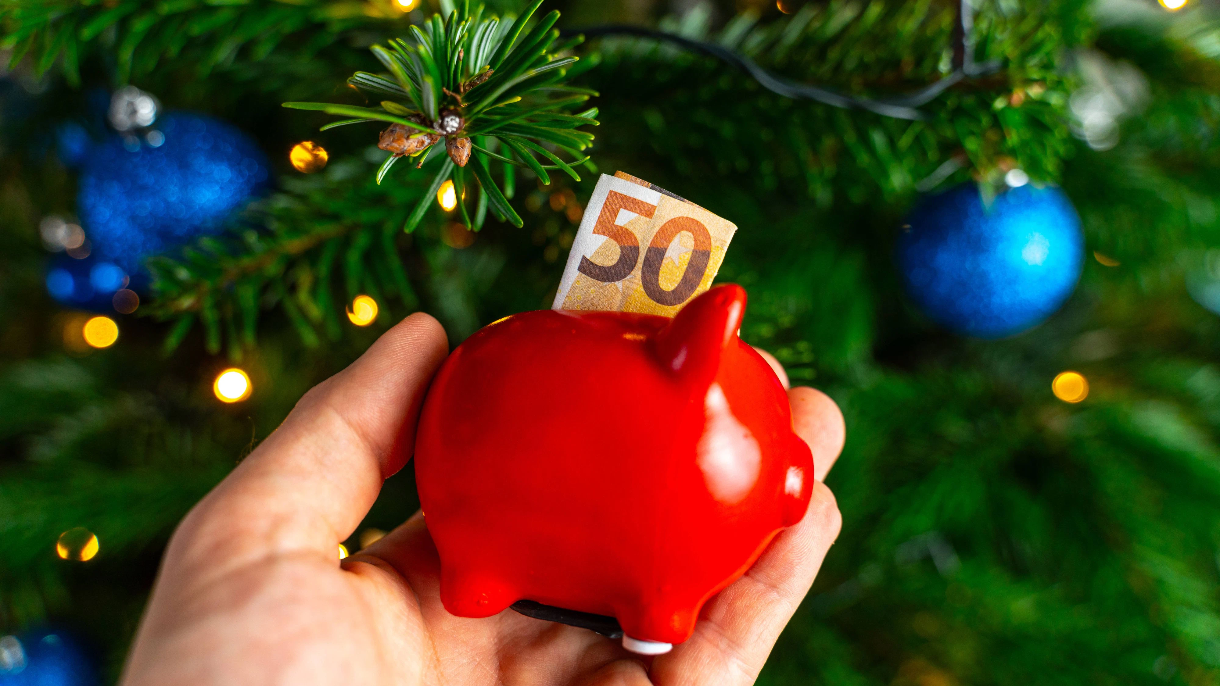 Augsburg, Bavaria, Germany - December 13, 2023: Hand holding a red piggy bank with a euro banknote in front of a decorated Christmas tree *** Hand hält rotes Sparschwein mit einem Euro Geldschein vor einen geschmückten Weihnachtsbaum