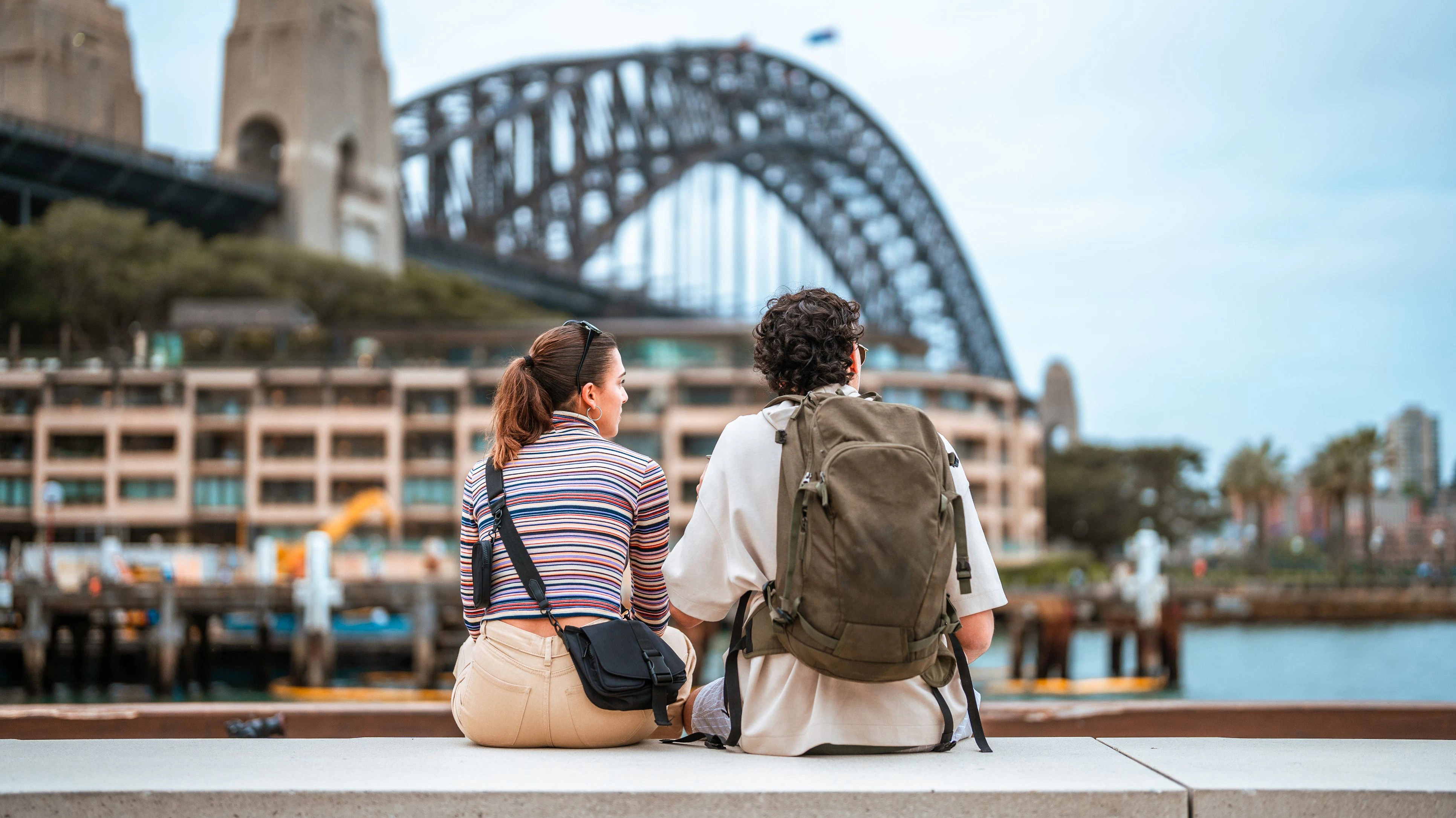 Candid portrait of young Hispanic couple sitting on harbor wall, wearing casual attire with backpacks, viewing Sydney Harbour Bridge under bright blue skies.