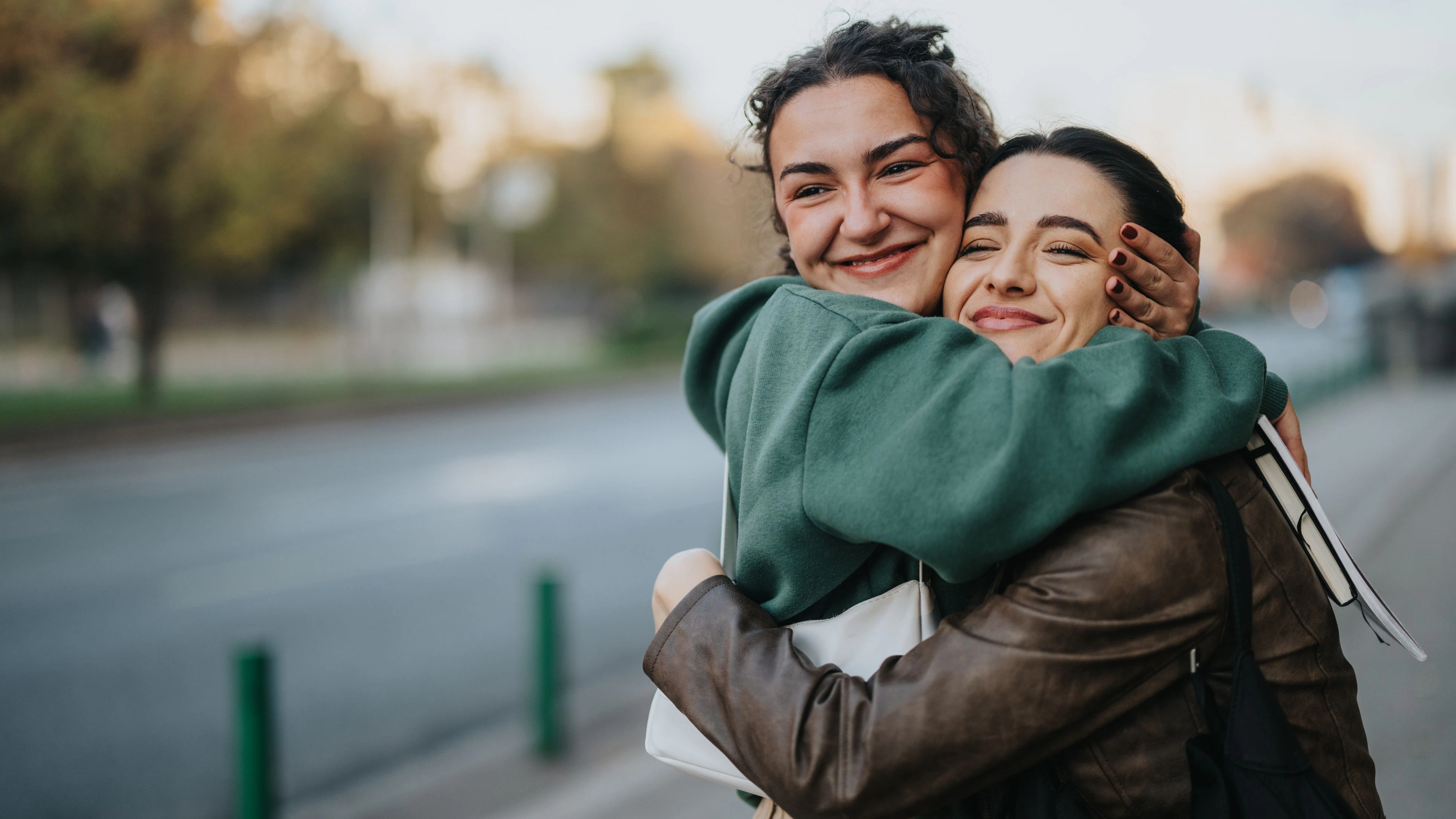 Two young women on a city sidewalk share a heartfelt embrace, smiling with genuine happiness. The casual outfits and urban backdrop highlight friendship, support, and everyday moments of joy.