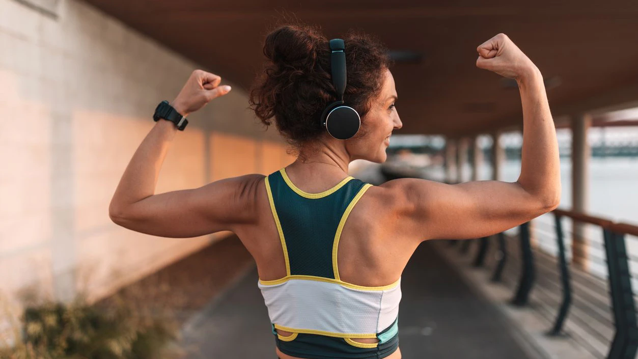 A confident woman flexes her muscles in an outdoor setting, showcasing her strength and dedication to fitness while enjoying a morning workout near the water.