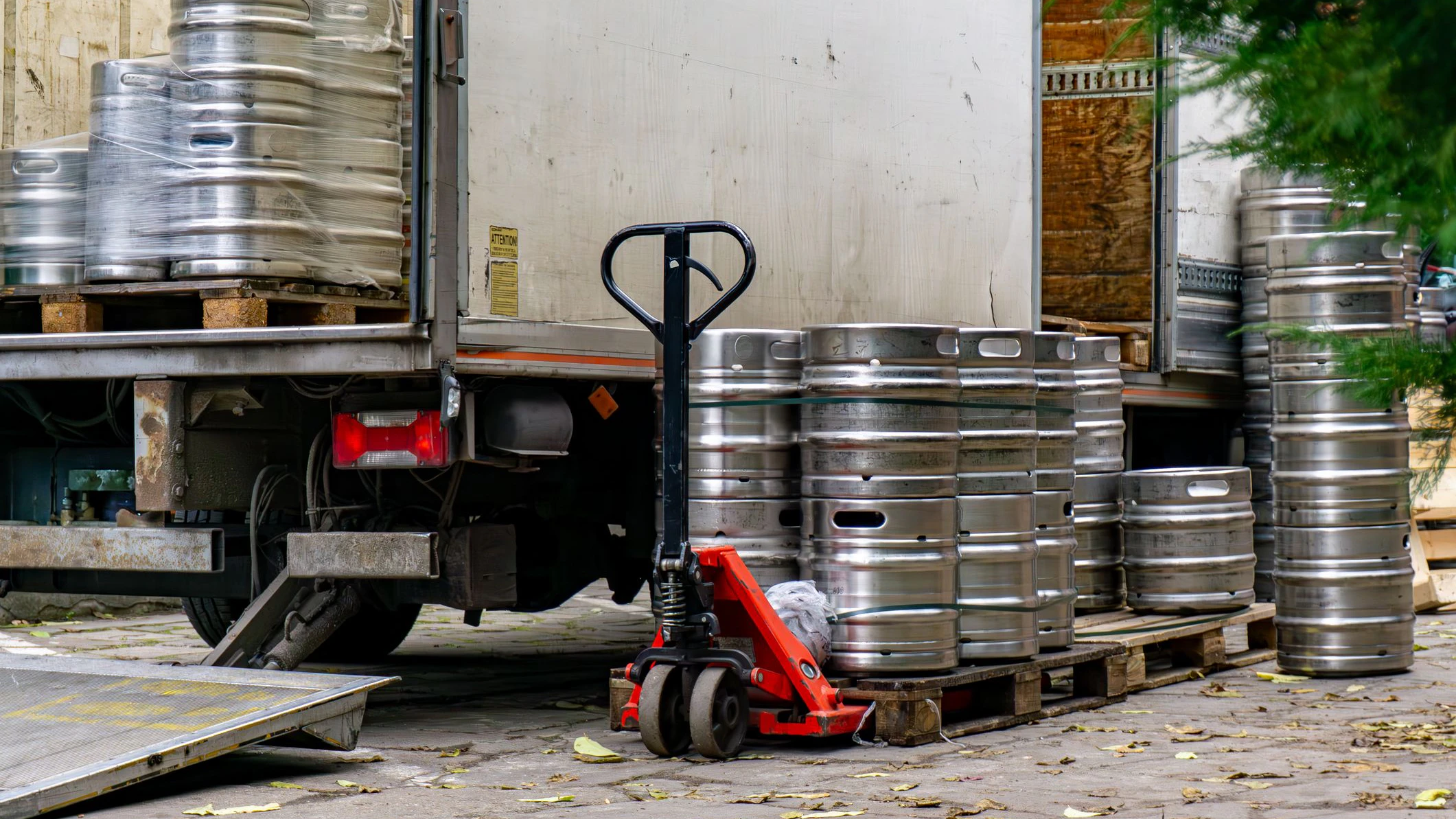 Entladen von Bierfässern aus einem Lieferwagen, roter Hubwagen neben Metallfässern, Herbstlaub auf dem Bürgersteig, Lagerbereich eines Restaurants in städtischer Umgebung.