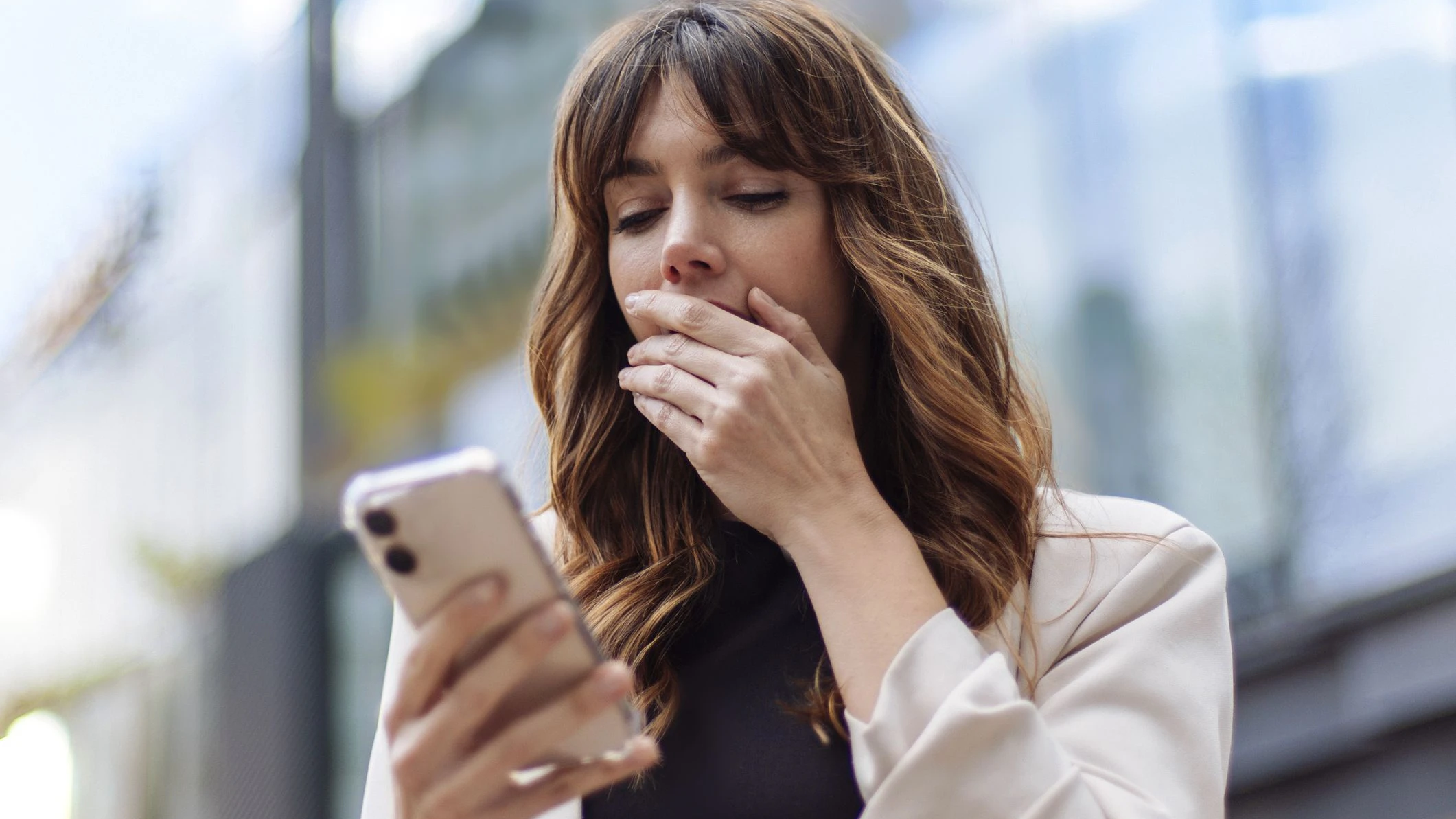 Eine junge Frau in einem Blazer reagiert überrascht und besorgt, während sie im Freien in einer städtischen Umgebung eine Nachricht auf ihrem Smartphone liest.
