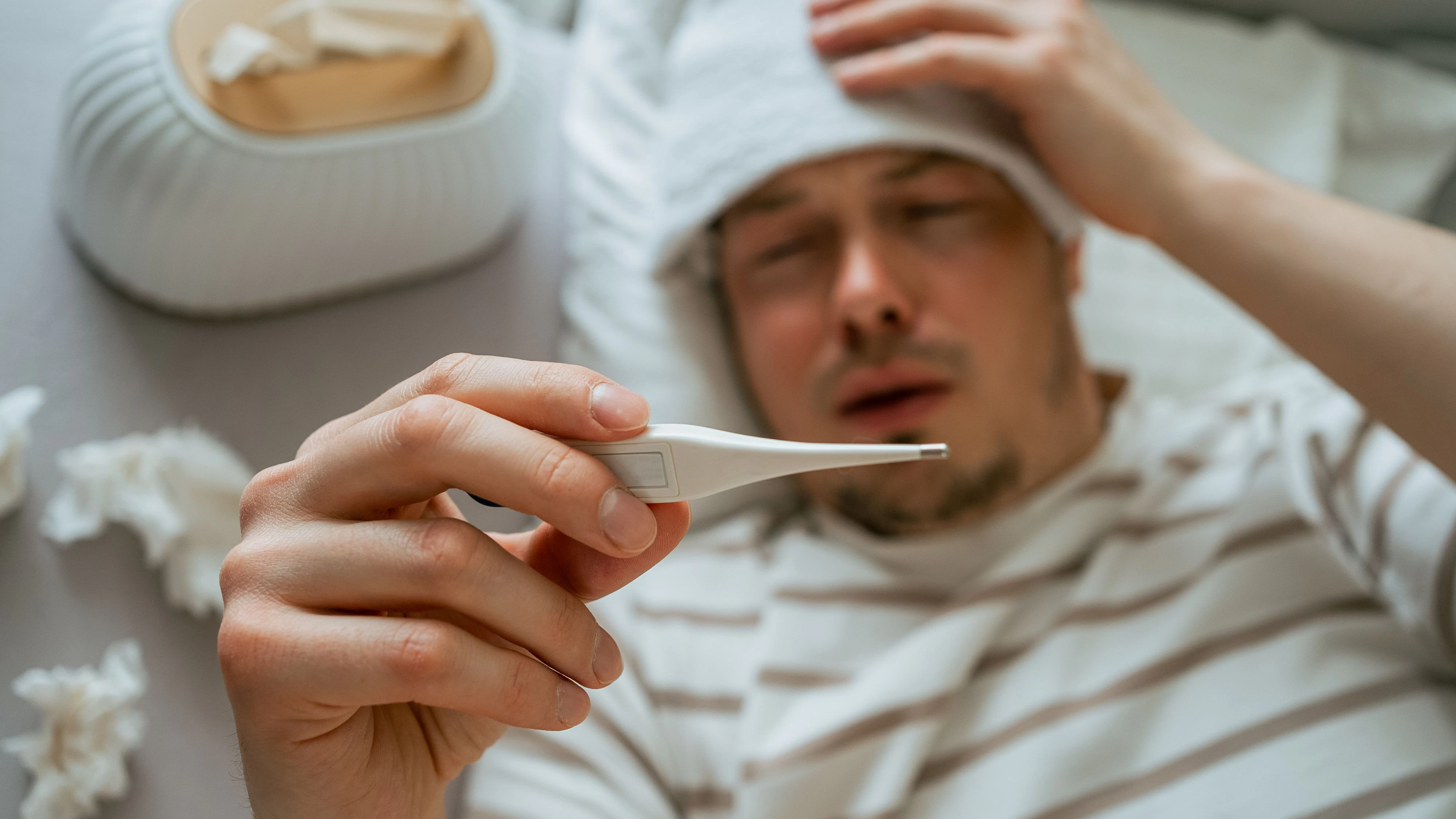 Sick man suffering from cold or flu, lying in bed with wet towel on his forehead, checking his body temperature with thermometer, tissues box and used tissues on the bed