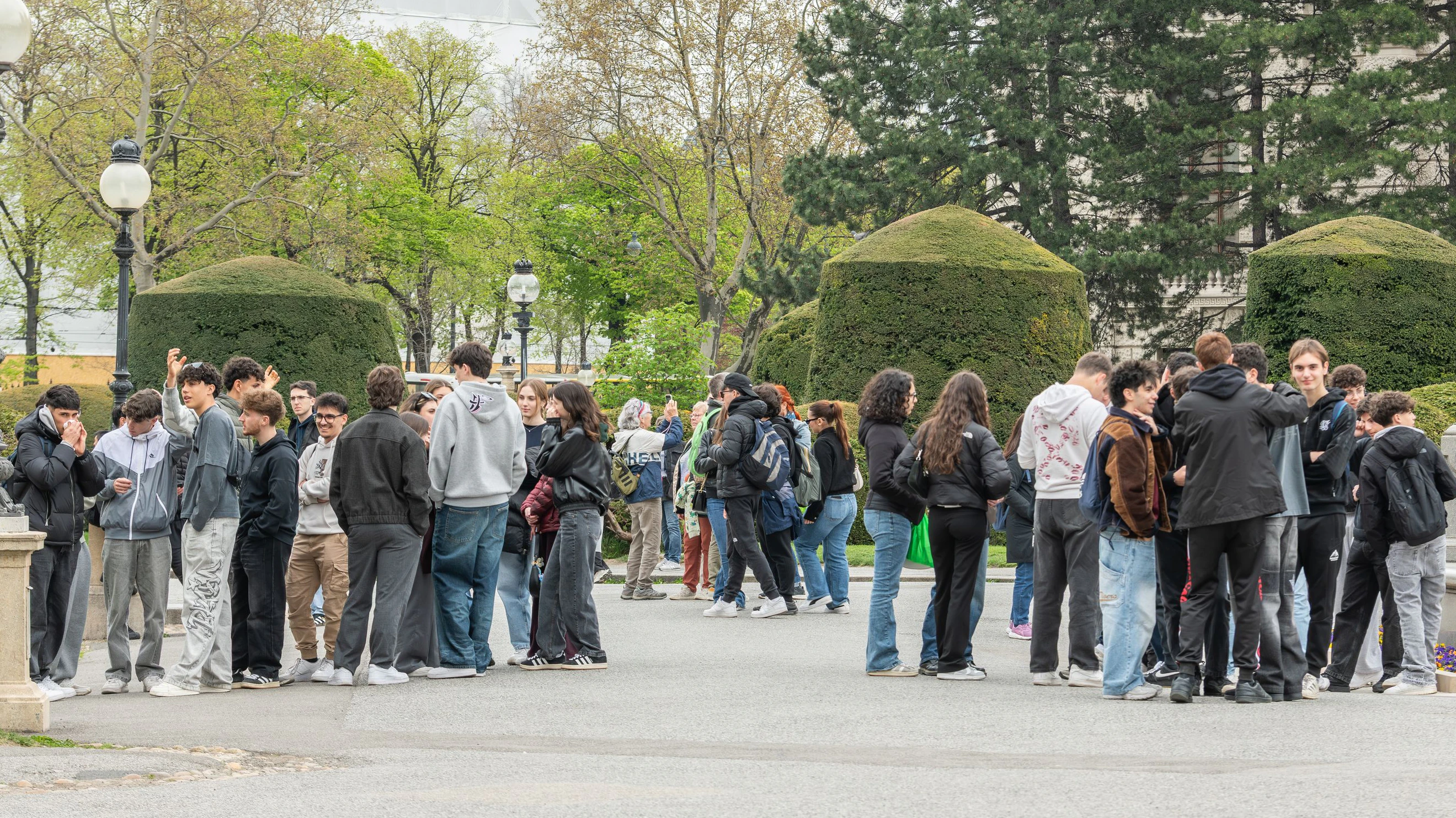 Maria-Theresien-Platz, Vienna, Austria. A large group of students gathers in a public square near the Maria Theresa monument, standing in small clusters and interacting. The scene reflects a school trip or group excursion in a historic urban park.