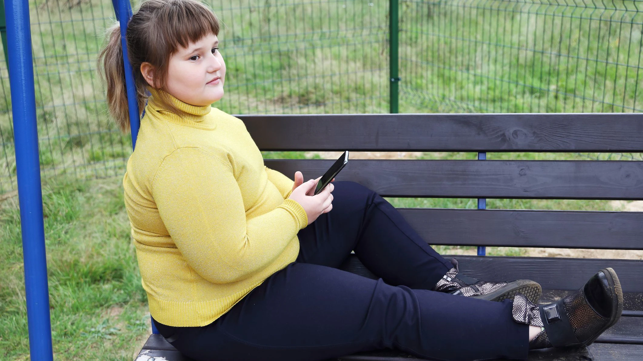 Plump thoughtful girl in yellow sweater sits alone on bench of swing outdoors, hold smartphone, looks aside and thinks